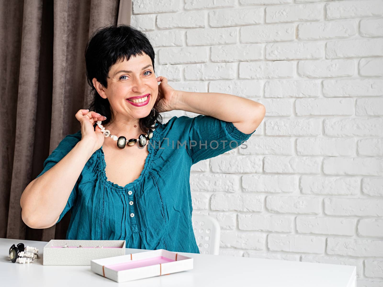 Beautiful smiling middle aged woman trying on the jewelery necklace sitting at the table with copy space