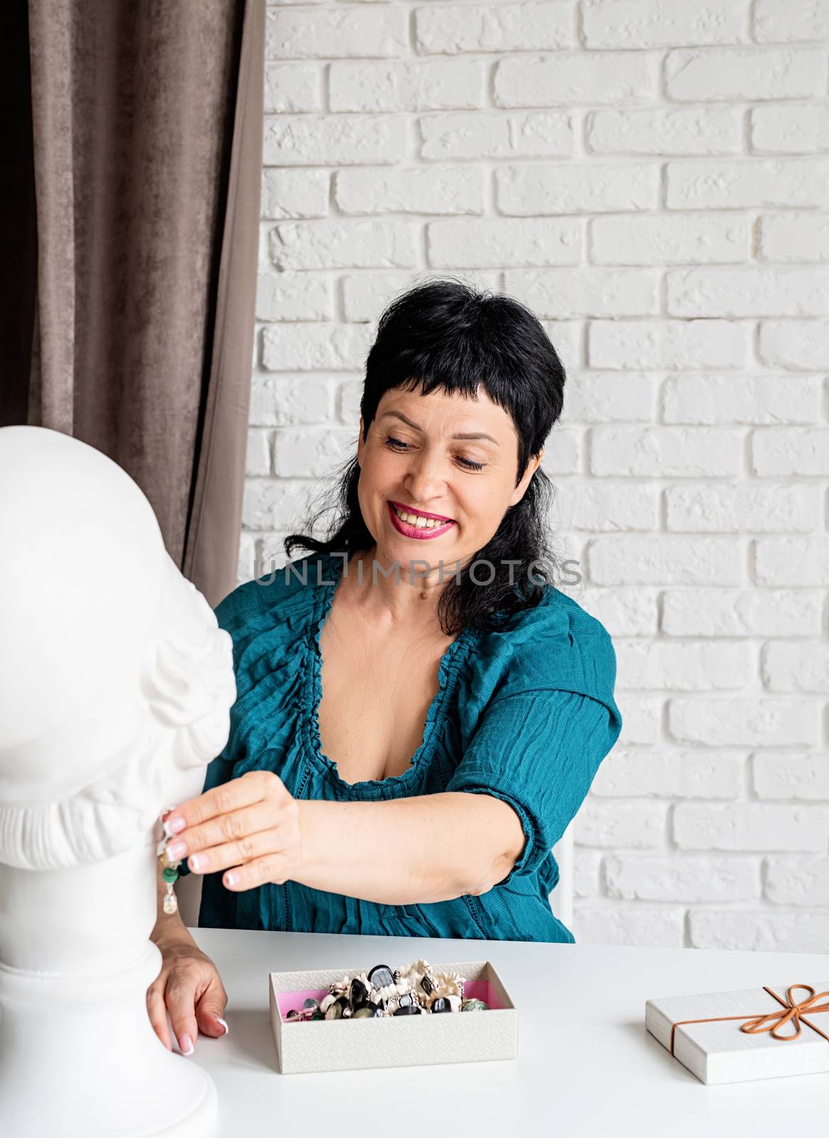 Beautiful smiling middle aged woman trying on the jewelery on the mannequin sitting at the table