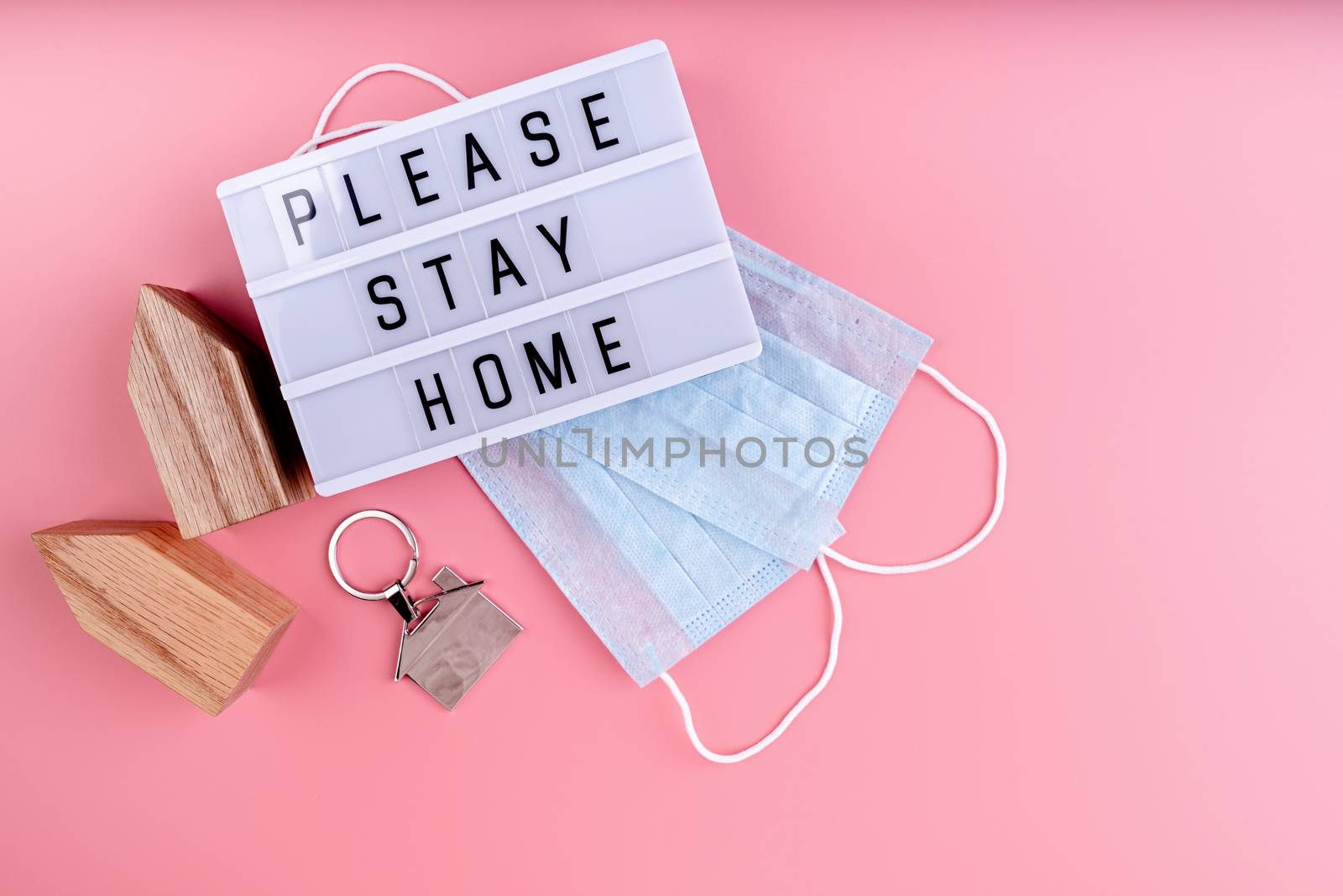 Stay home. Self quarantine. The words Please Stay Home on a lightbox with wooden house and masks top view on pink background