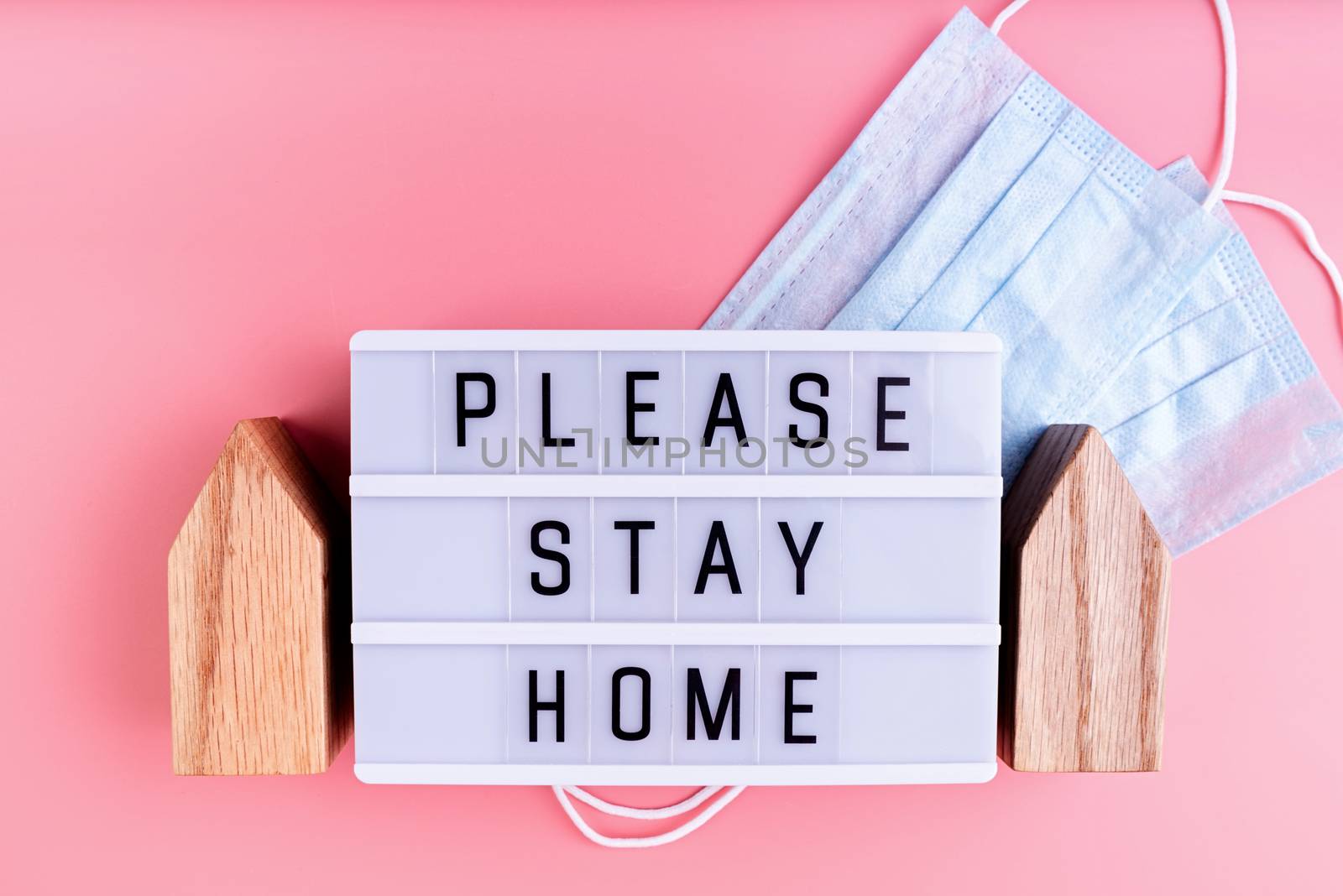 Stay home. Self quarantine. The words Please Stay Home on a lightbox with wooden house and masks top view on pink background