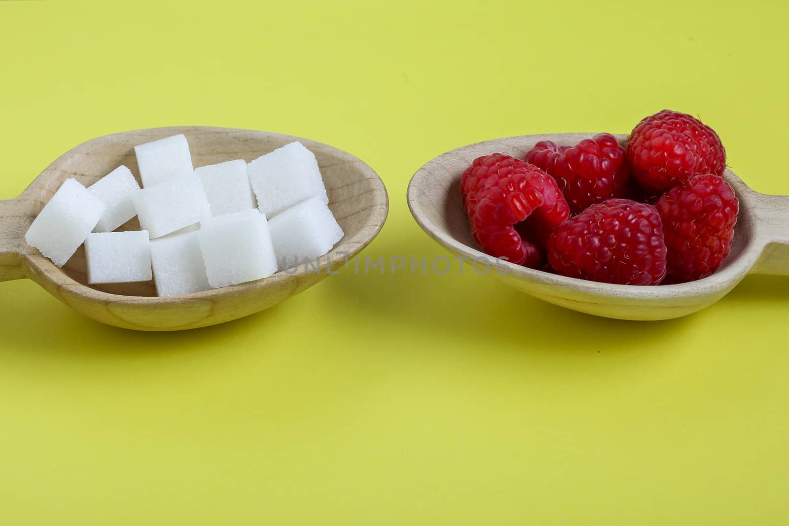 Healthy raspberries and unhealthy sugar in wooden spoons with yellow background