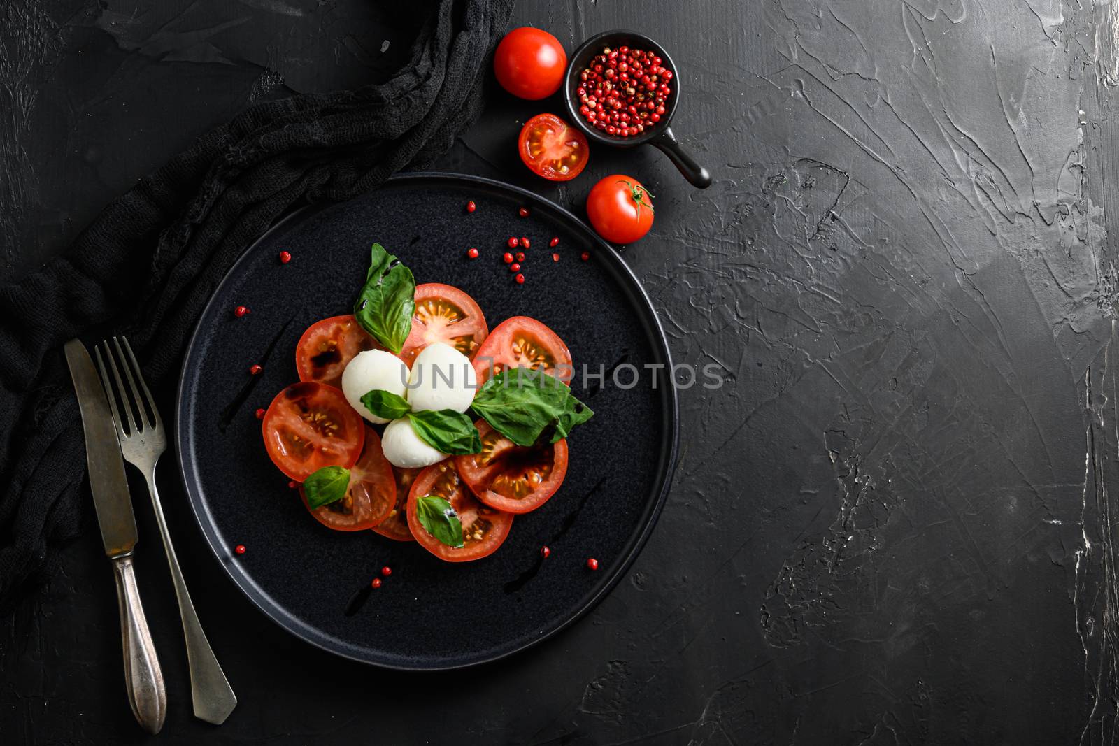 Caprese salad Italian cuisine concept Tomato and mozzarella slices with basil leaves on black ceramic platwantipasta black textured background close up with fork and knife space for text by Ilianesolenyi