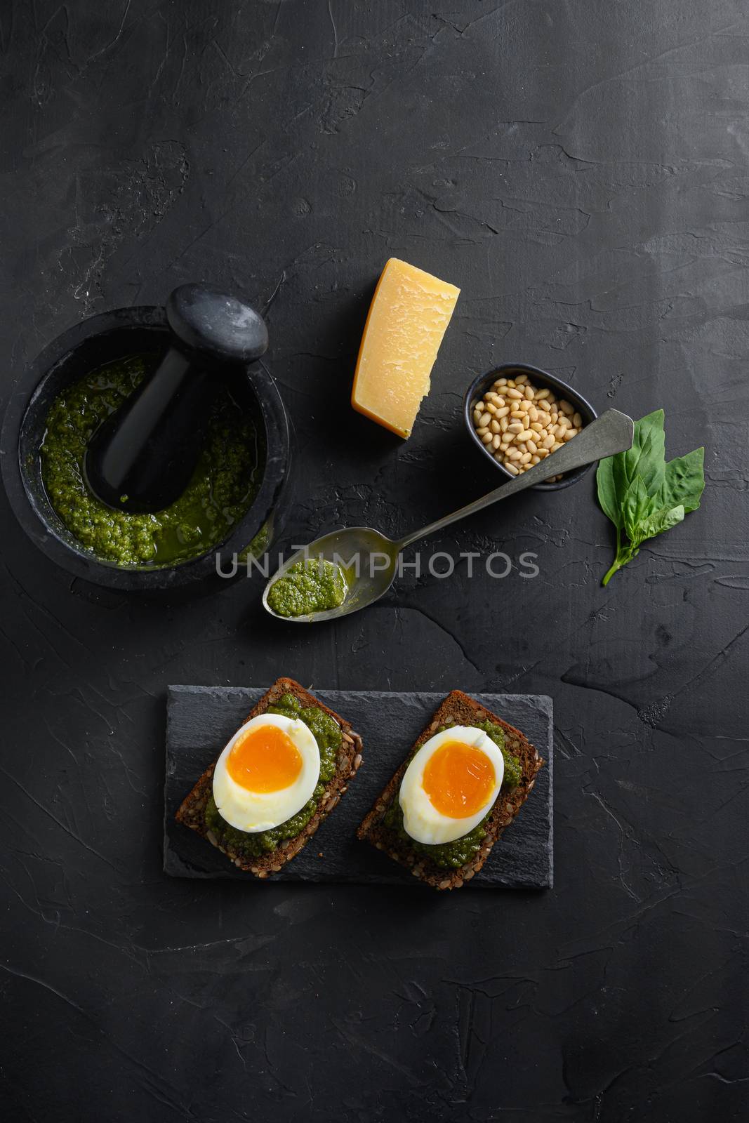 Fresh Green basil pesto in mortar bowl homemade on old rustic wood eggs panini bread with pesto silver spoon on black slateand hot sriracha sauce italian breakfast with ingredients green pesto top view on black stone concrete table surface by Ilianesolenyi
