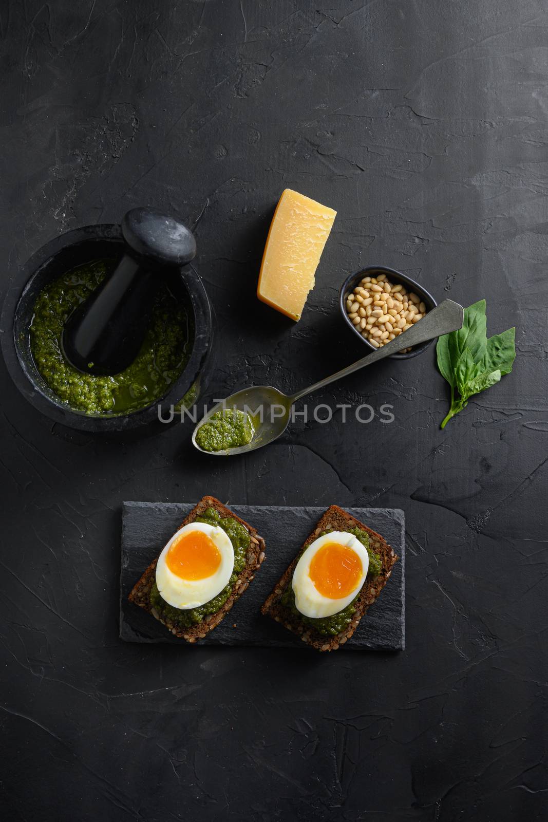 Fresh Green basil pesto mortar bowl homemade on old rustic wood eggs panini bread with pesto silver spoon on black slateand hot sriracha sauce italian breakfast with ingredients green pesto top view on black stone concrete table surface.