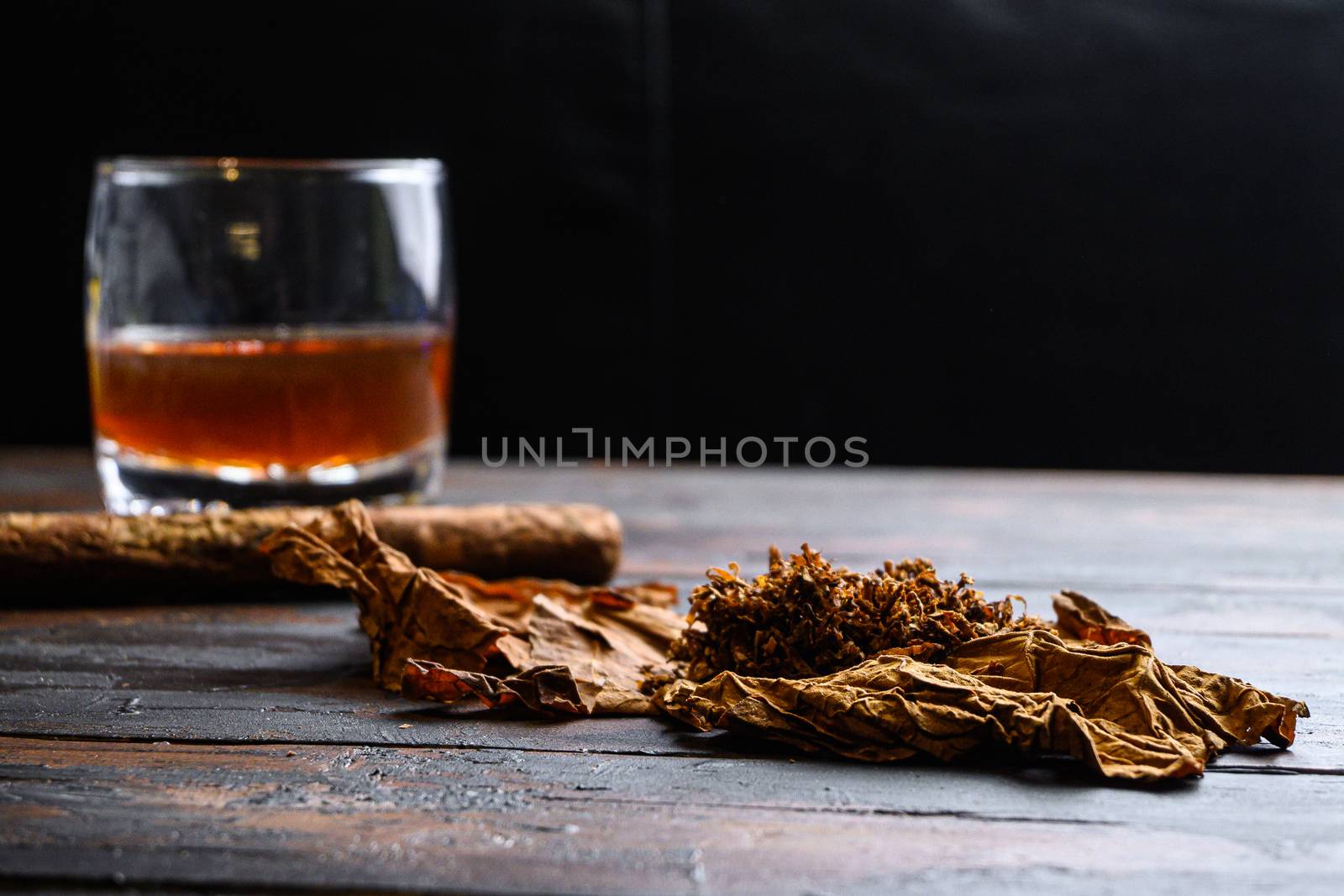 Dried tobacco leave and cut tobacco with cigar and whiskey rum on wood background on vintage dark table. side view space for text.