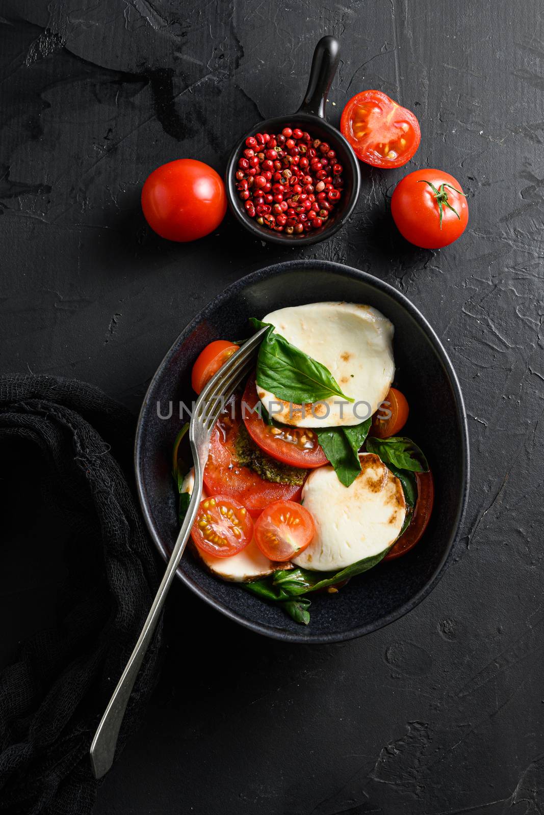 Cherry tomatoes, mozzarella buffalo cheese, basil, pesto sauce, and spices on black slate stone chalkboard. Italian traditional caprese salad ingredients. Mediterranean food in black bowl with fork.