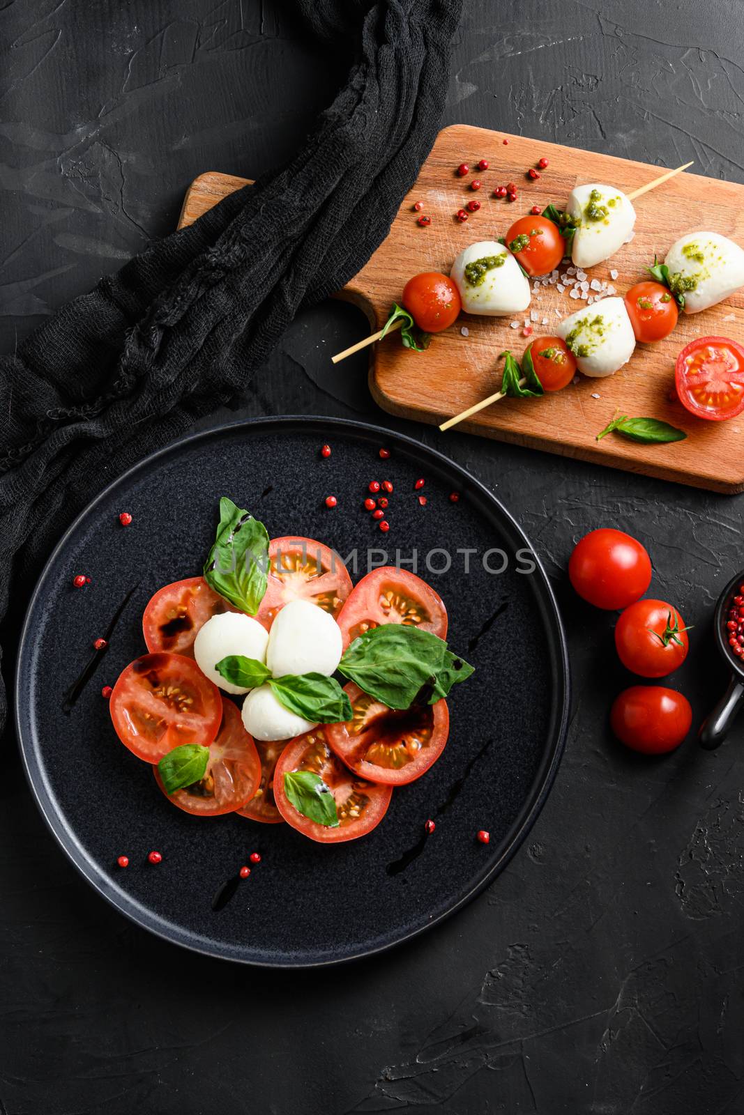 Caprese salad Tomato and mozzarella slices with basil leaves on sticks skewer and on black ceramic platwantipasta black textured background top view.