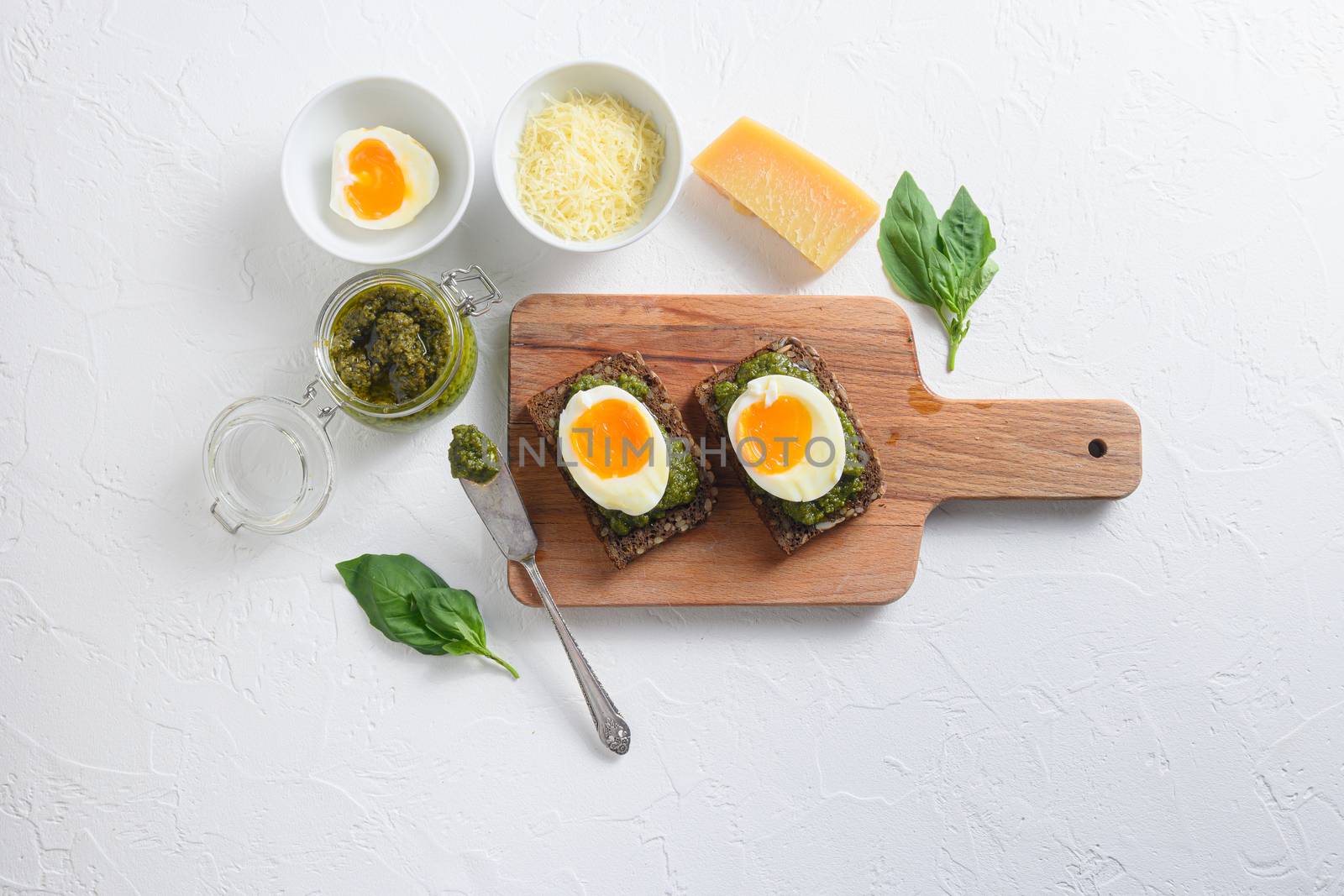 Fresh Green basil pesto in glass jar homemade on old rustic wood eggs panini bread with pesto knife and hot sriracha sauce italian breakfast with ingredients green pesto top view on white concrete table by Ilianesolenyi