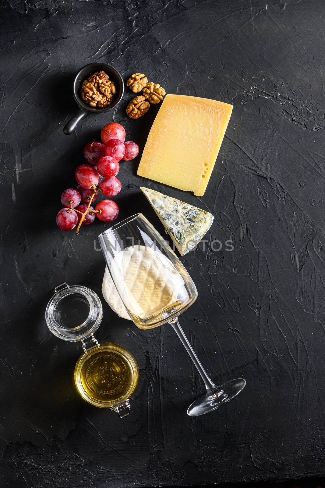Cheese appetizer selection or whine snack set. Variety of mold french cheese, grapes, pecan nuts, white sauvignon wine and honey over black backdrop, top view, copy space vertical by Ilianesolenyi