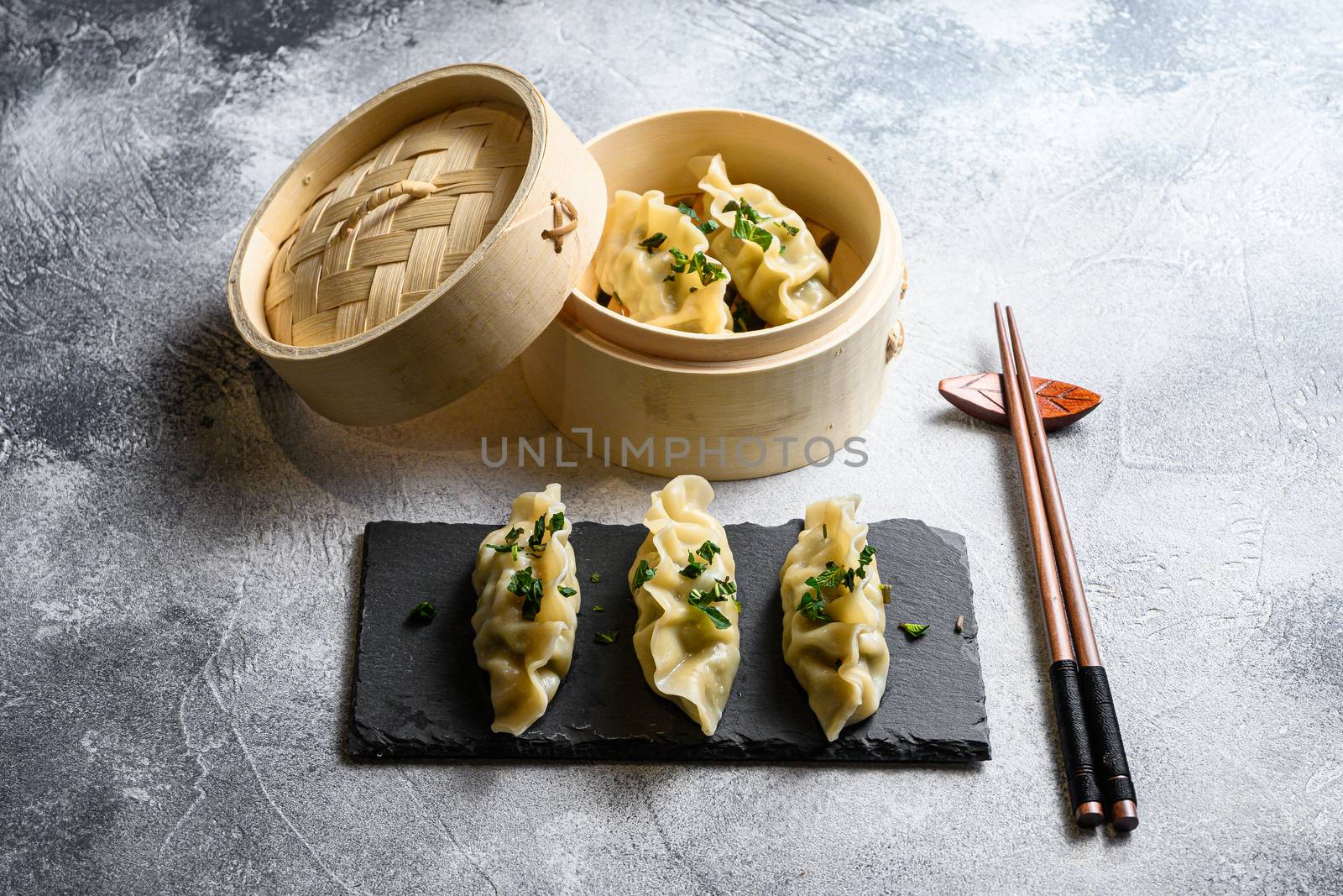 Dim sum dumplings in steamer and on a stone plate with chopsticks and fresh herbs side view.