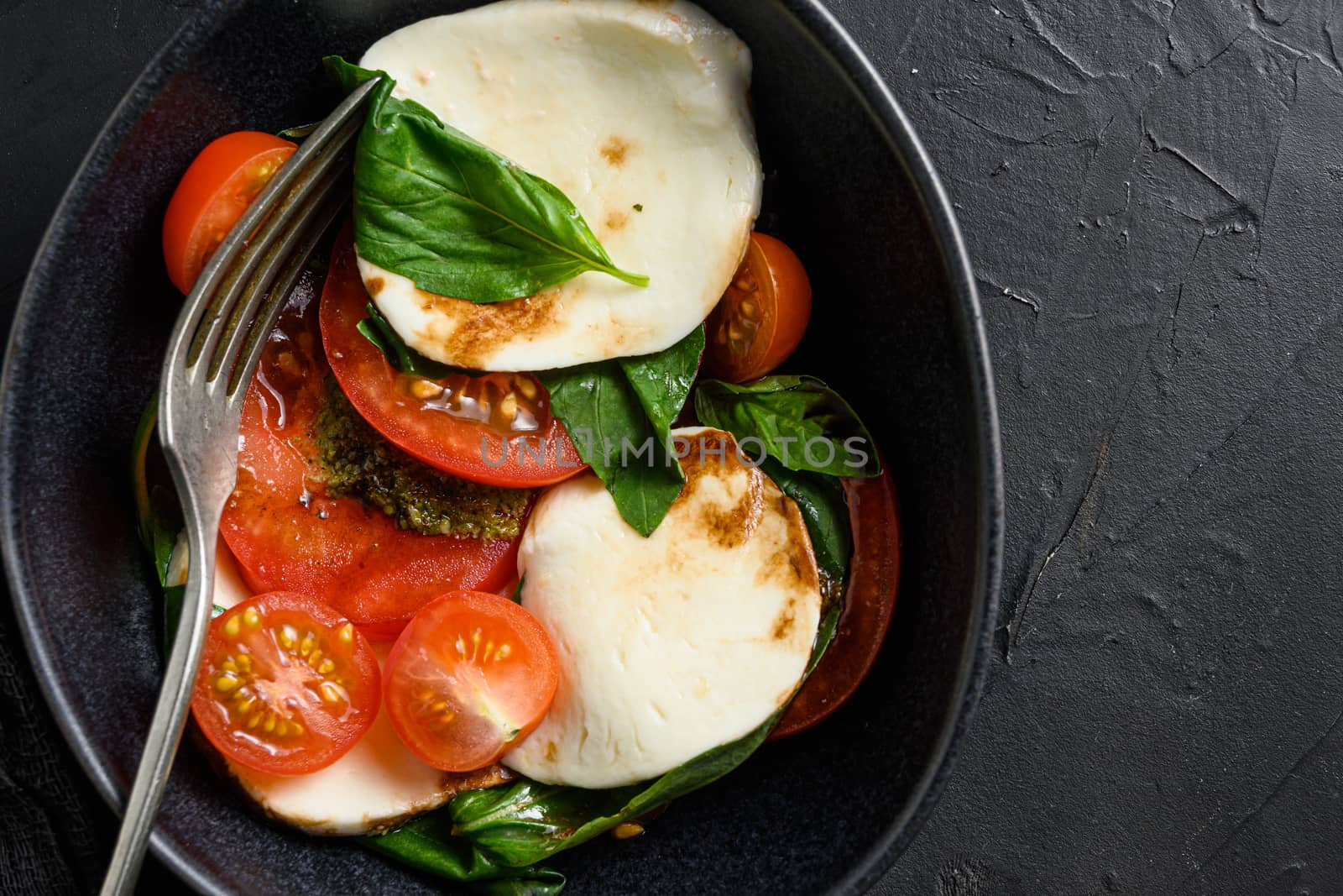 Cherry tomatoes, mozzarella buffalo cheese, basil, pesto sauce, and spices on black slate stone chalkboard. Italian traditional caprese salad ingredients. Mediterranean food in black bowl with fork close up space for text.