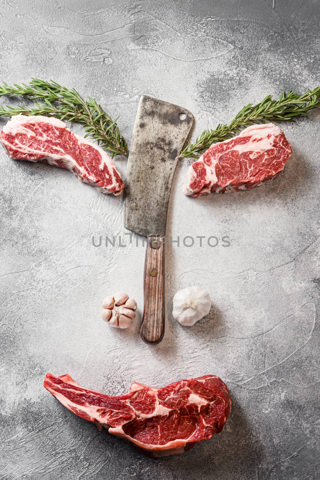 Concept Various beef black angus beauty steak or Cowboy steak, with chuck eye roll cut with the meat american cleaver knife over grey stone table slate cutting top view face styled art with seasonings, tomatoes, garlic, rosemary by Ilianesolenyi