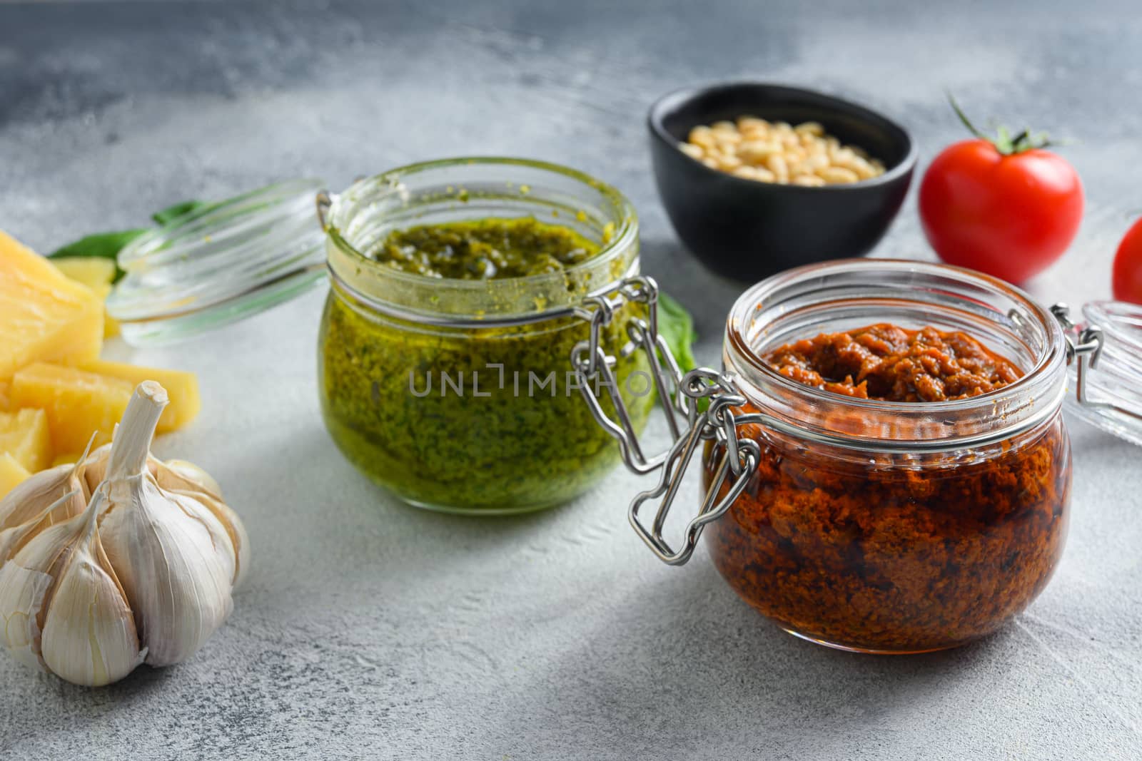 Home made Vegan pesto normal pesto and Red pesto and cooking ingredients Parmesan cheese, basil leaves, pine nuts, olive oil, garlic, salt, pepper side view on grey concrete surface close up.