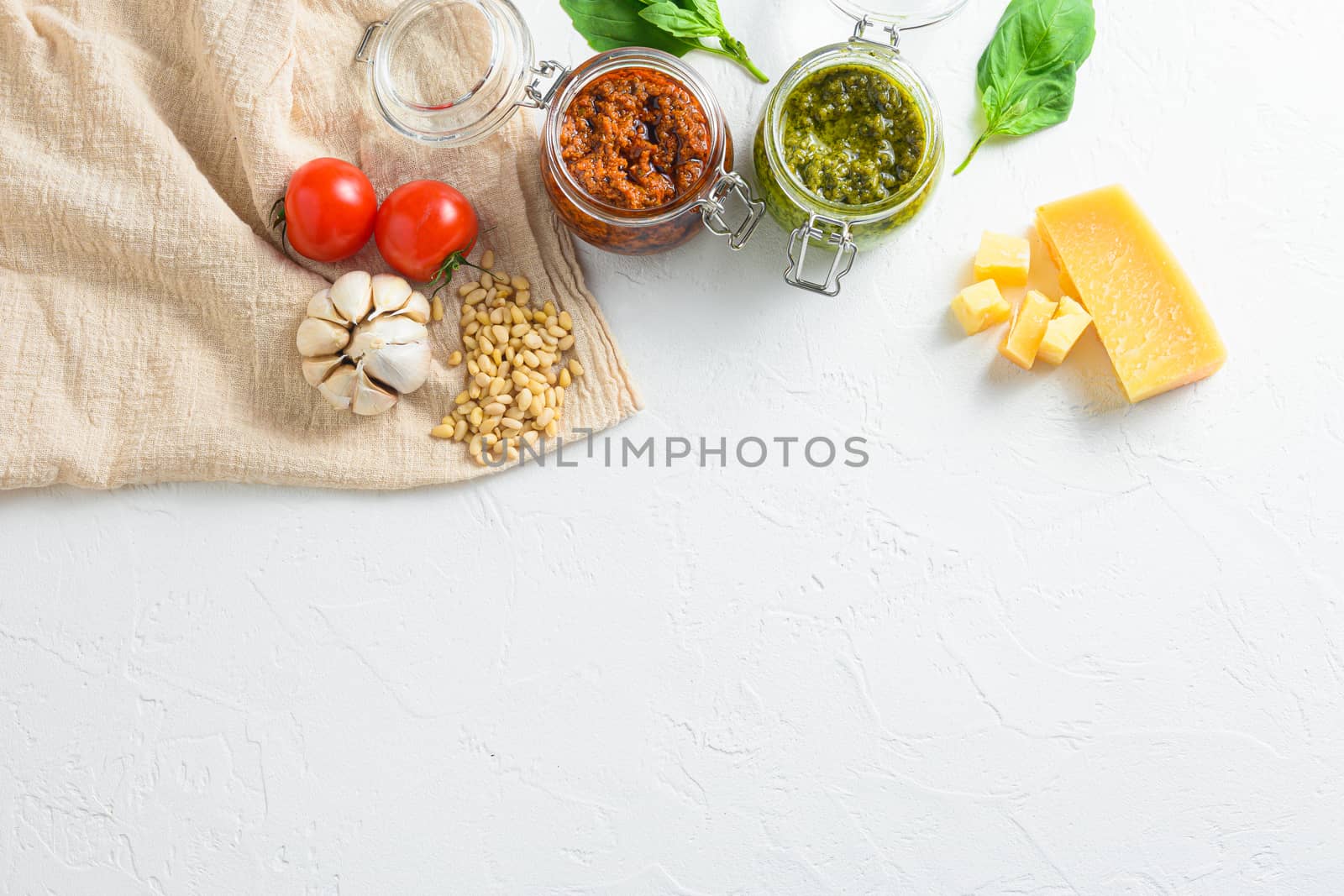 red pesto and green pesto Glass jars with cooking italian recipe ingredients Parmesan cheese, basil leaves, pine nuts, olive oil, garlic, salt, tomatoes top view on white textured concrete surface space for text