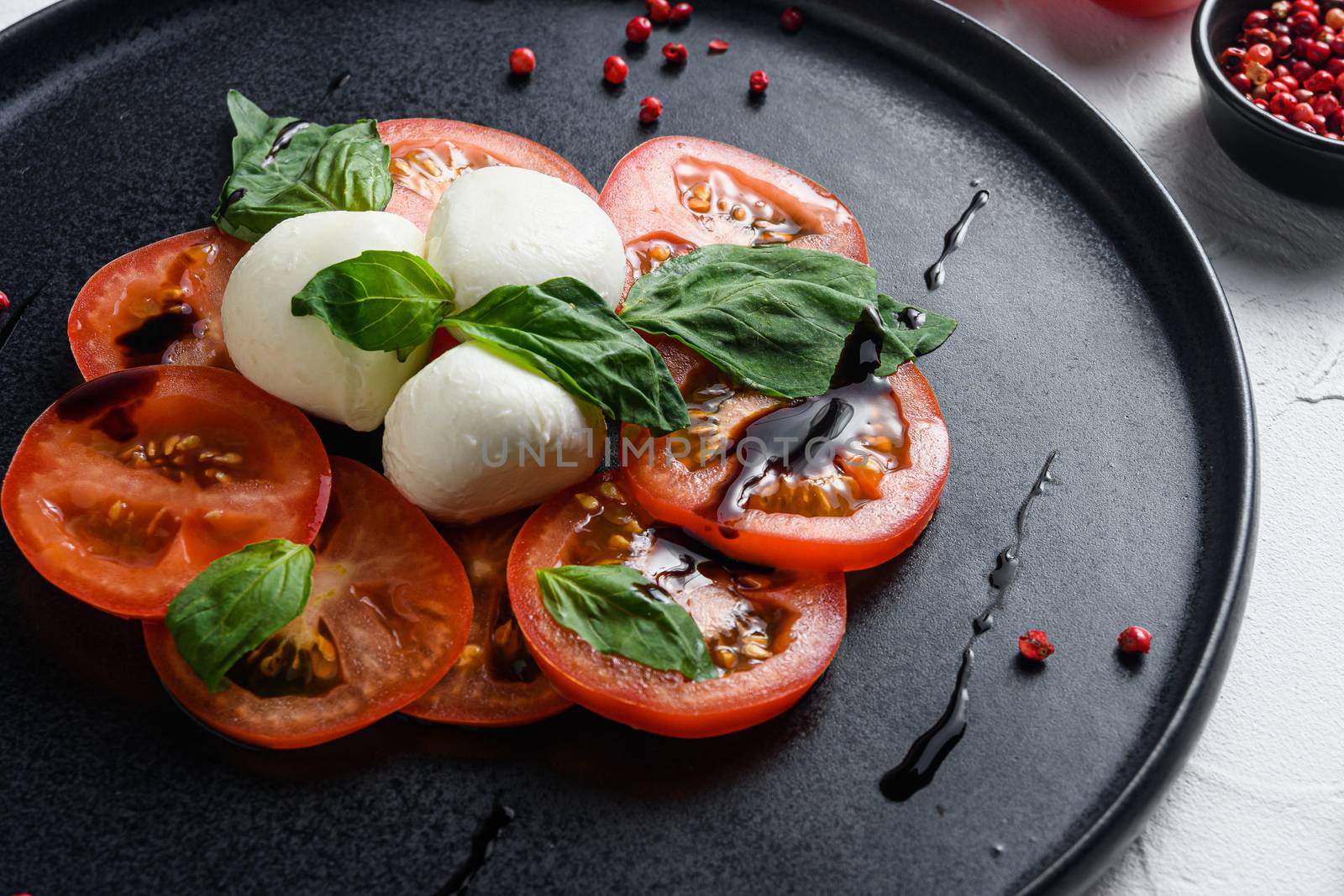 Caprese fresh italian salad with tomatoes, mozzarella, green basil on dark slate plate over white background close up selective focus.