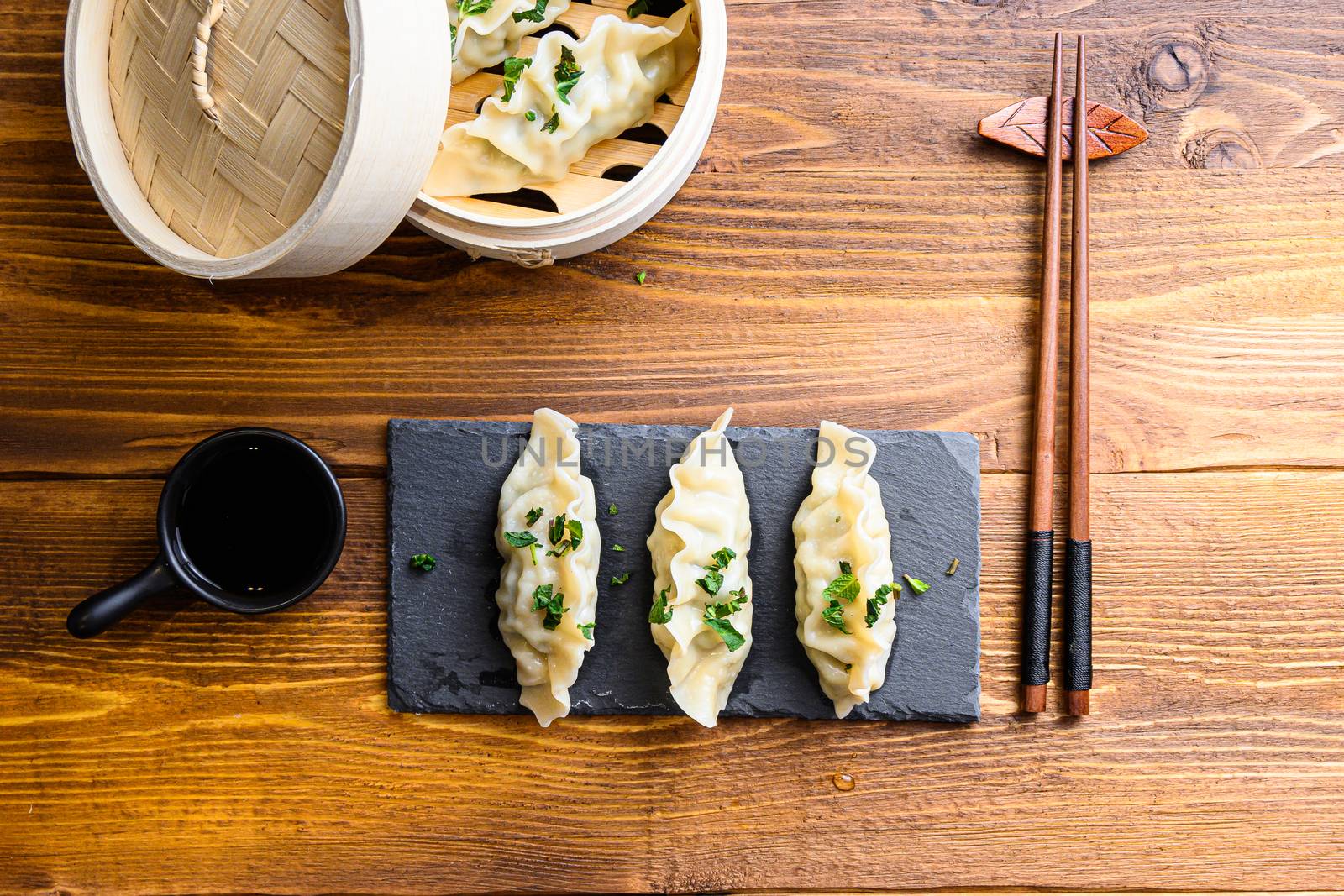 Asian dumplings on stone slate and in wooden steamer over traditional wood table with soy sauce and chopsticks and fresh herbs top view.