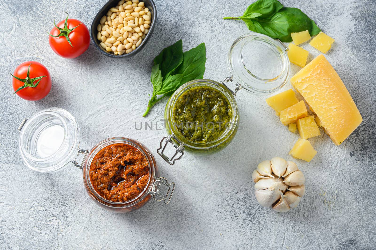 Glass jars with red and green pesto and cooking ingredients Parmesan cheese, basil leaves, pine nuts, olive oil, garlic, salt, pepper top view on grey concrete surface .