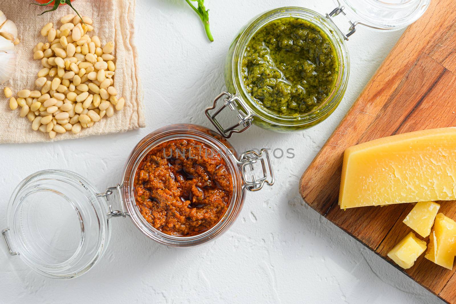 Glass jars with red and green pesto and cooking ingredients Parmesan cheese, basil leaves, pine nuts, white stone background, close up top view. by Ilianesolenyi