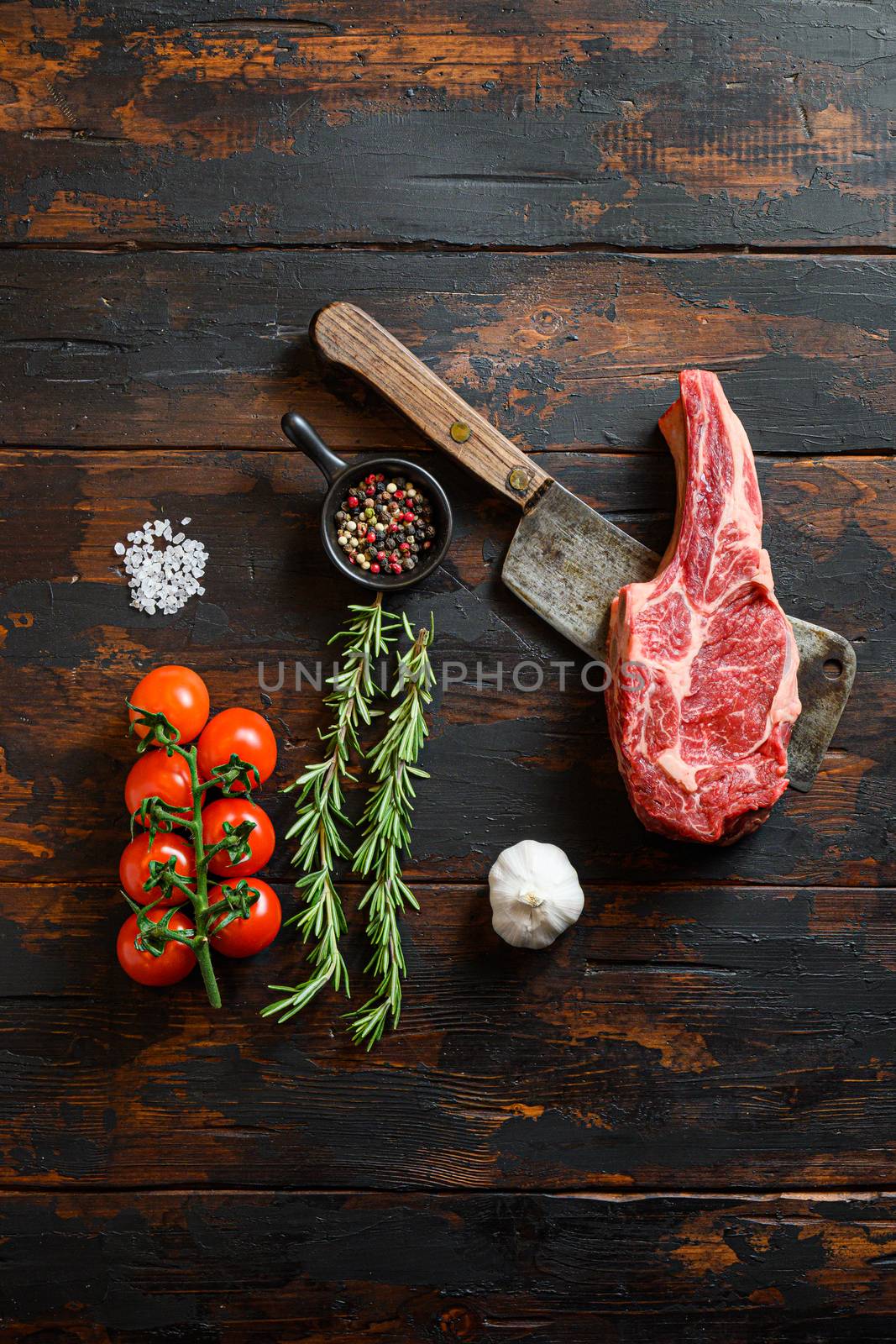 Fresh Cowboy steak cut on meat cleaver. Organic farm marbled prime black angus beef. Dark wooden background. Top view. With seasonings, peppercorns, chilie, rosemary,salt,garlic. Copy space. Vertical