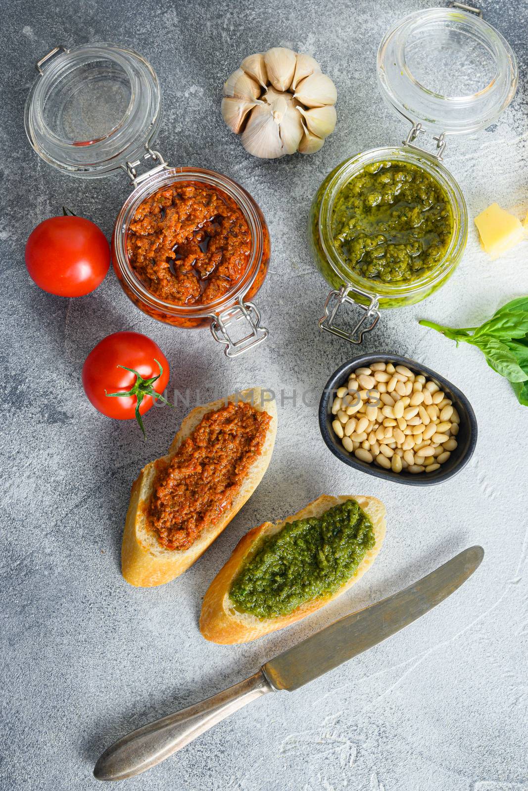 Glass jars with red and green pesto and cooking italian recipe ingredients Parmesan cheese, basil leaves, pine nuts, olive oil, garlic, salt, tomatoes breakfast panini with sauce top view on grey concrete surface .