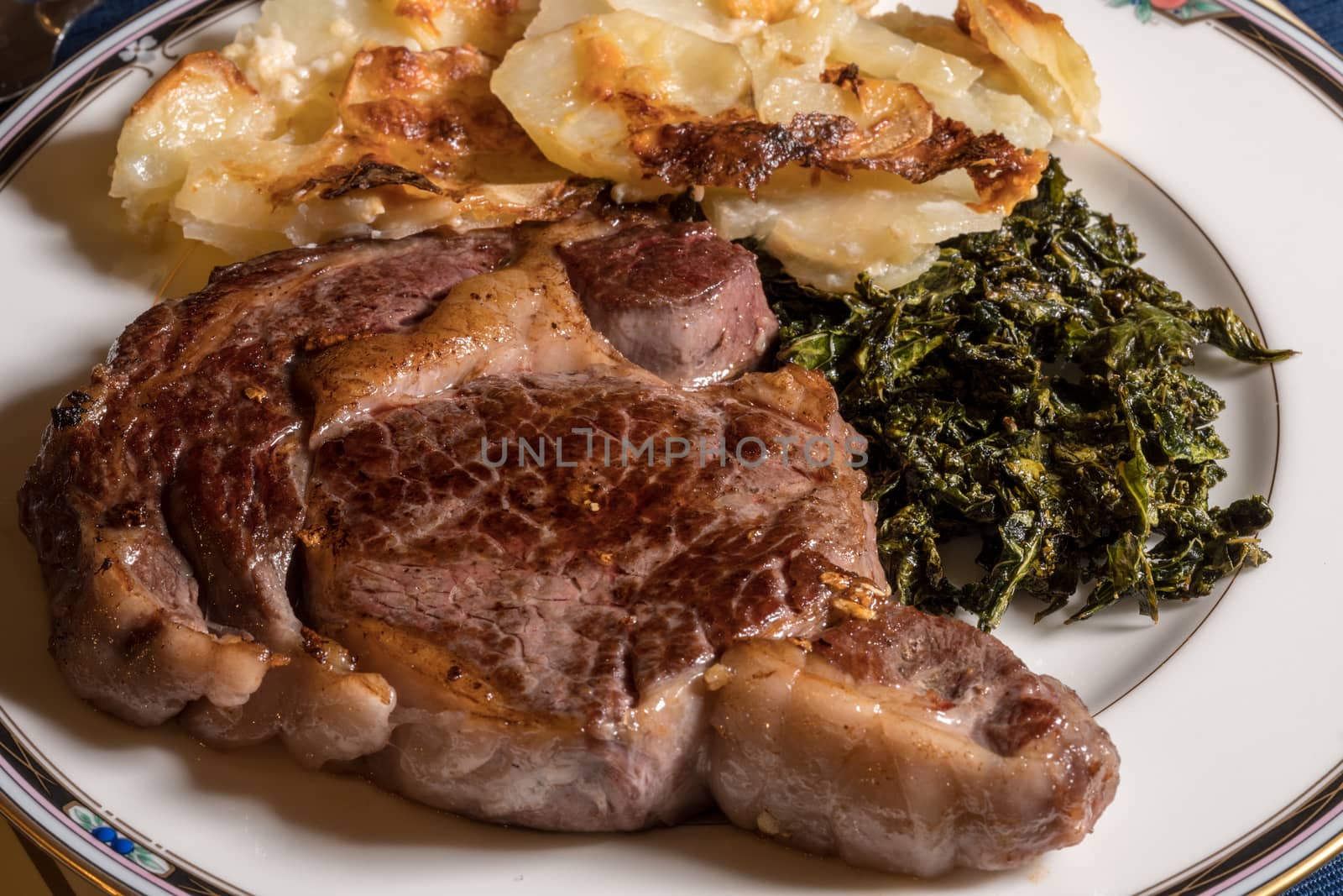 Large grilled ribeye steak on white plate with roasted kale and sliced potatoes