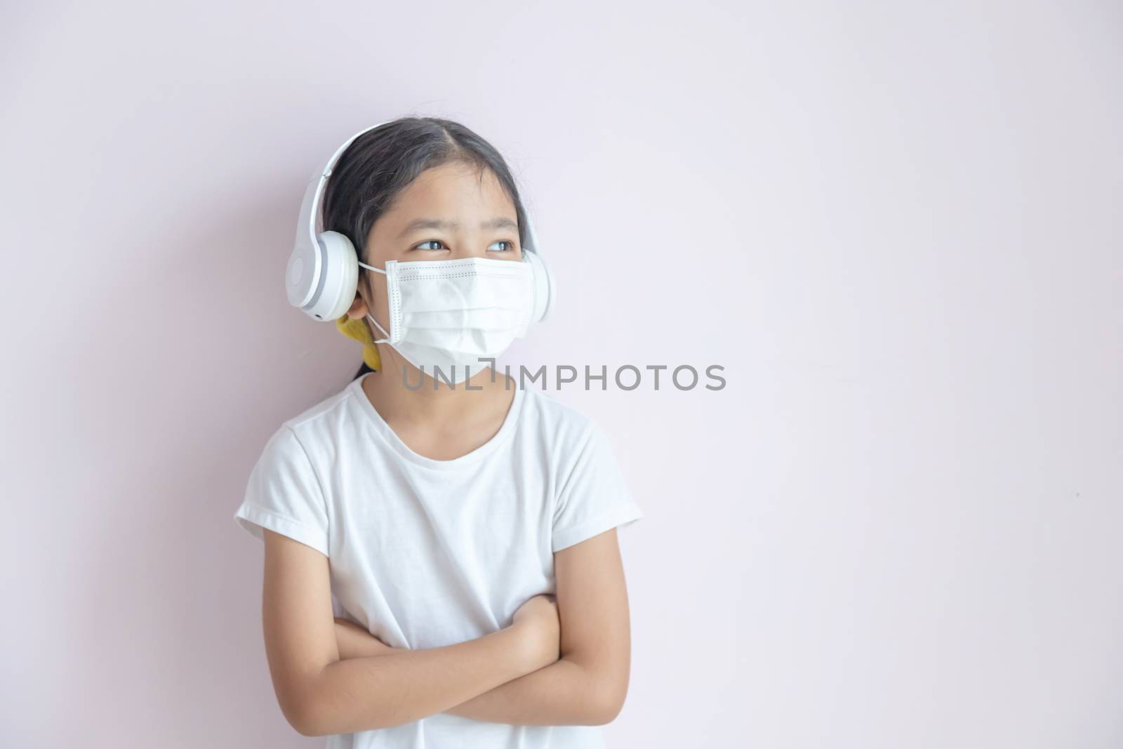 A little Asian girl wearing a medical protective mask and earphones. The child girl standing cross one's arm on the color background. The epidemic of the flu, Coronavirus or COVID-19 and illness concept.