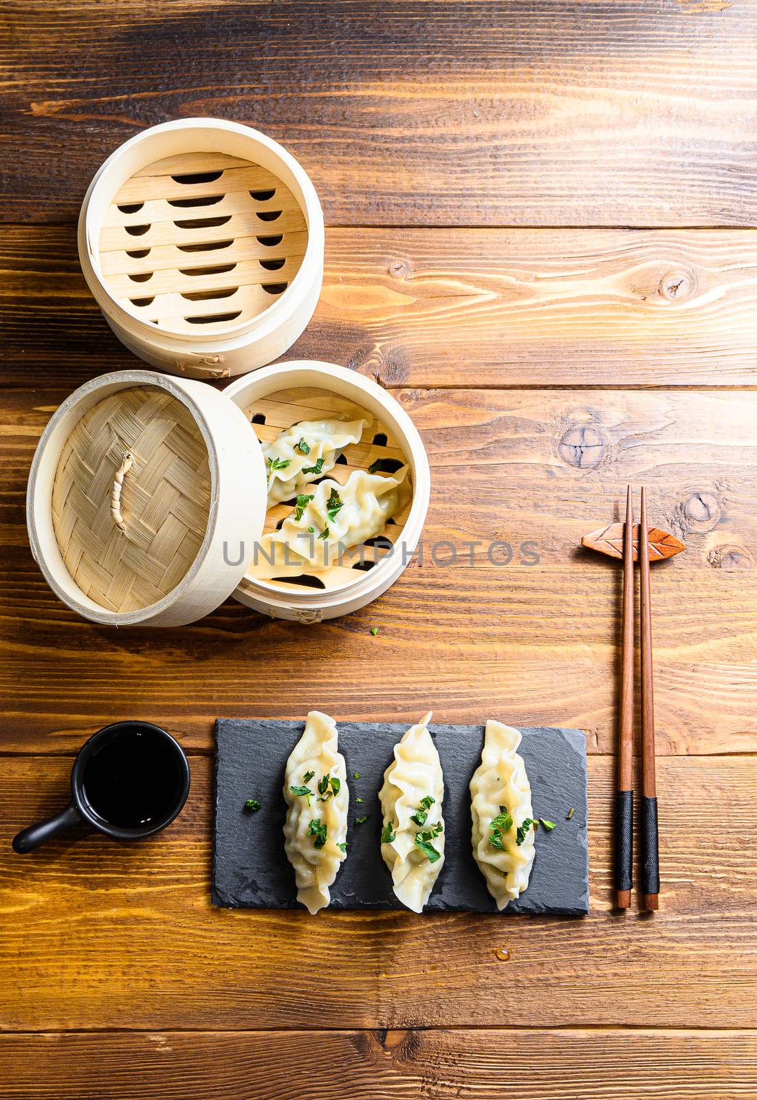 Asian dumplings on stone slate and in wooden steamer over traditional wood table with soy sauce and chopsticks and fresh herbs top view space for text by Ilianesolenyi
