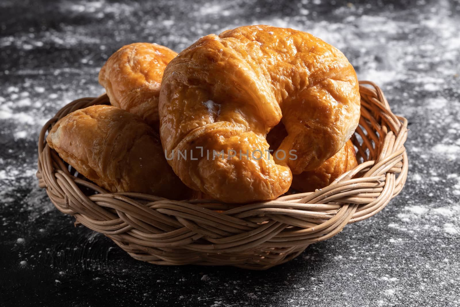 Croissants in a wicker basket placed on a black floor by Nikkikii