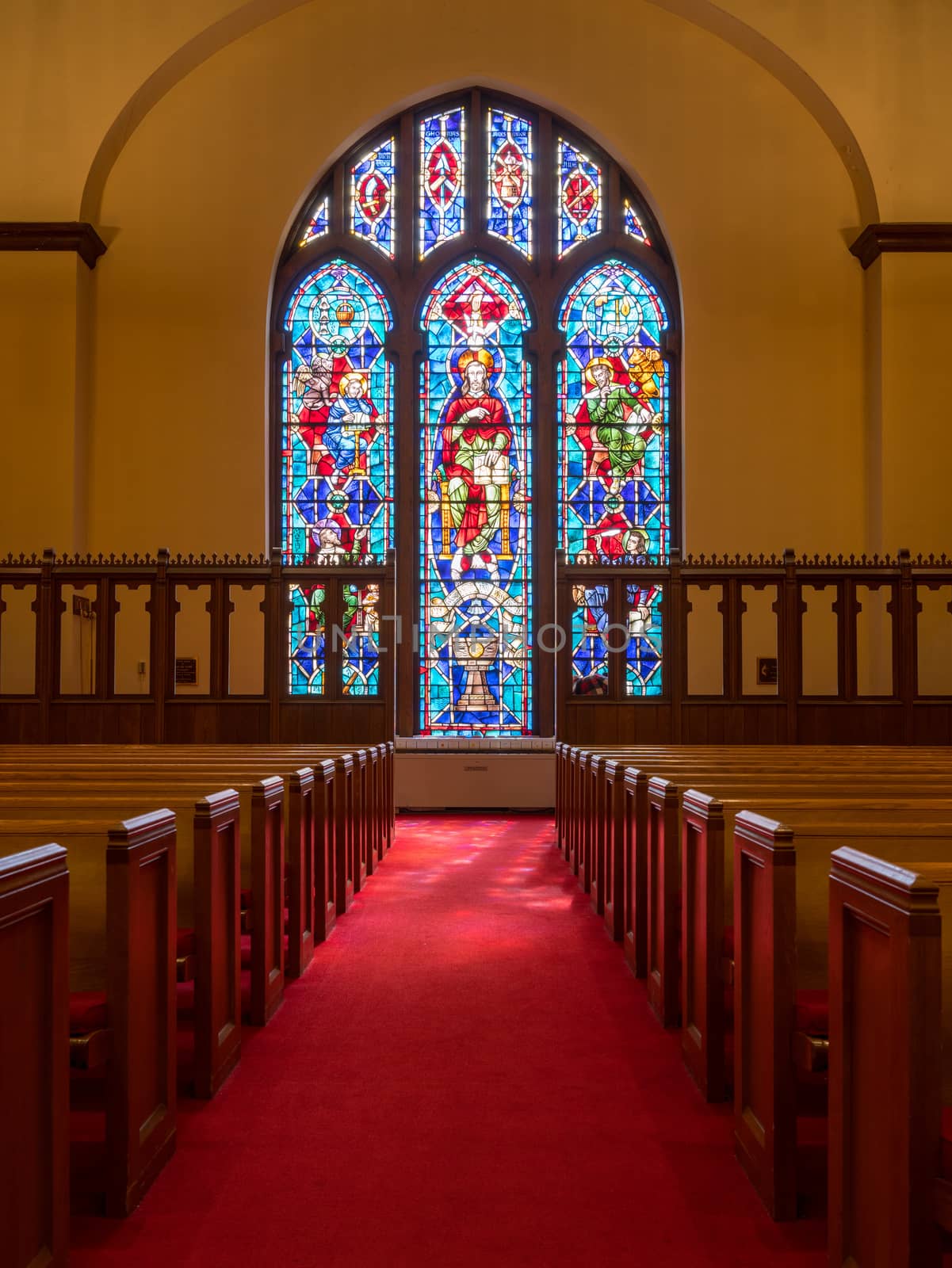 Red carpeted church aisle between pews illuminated by light from stained glass window