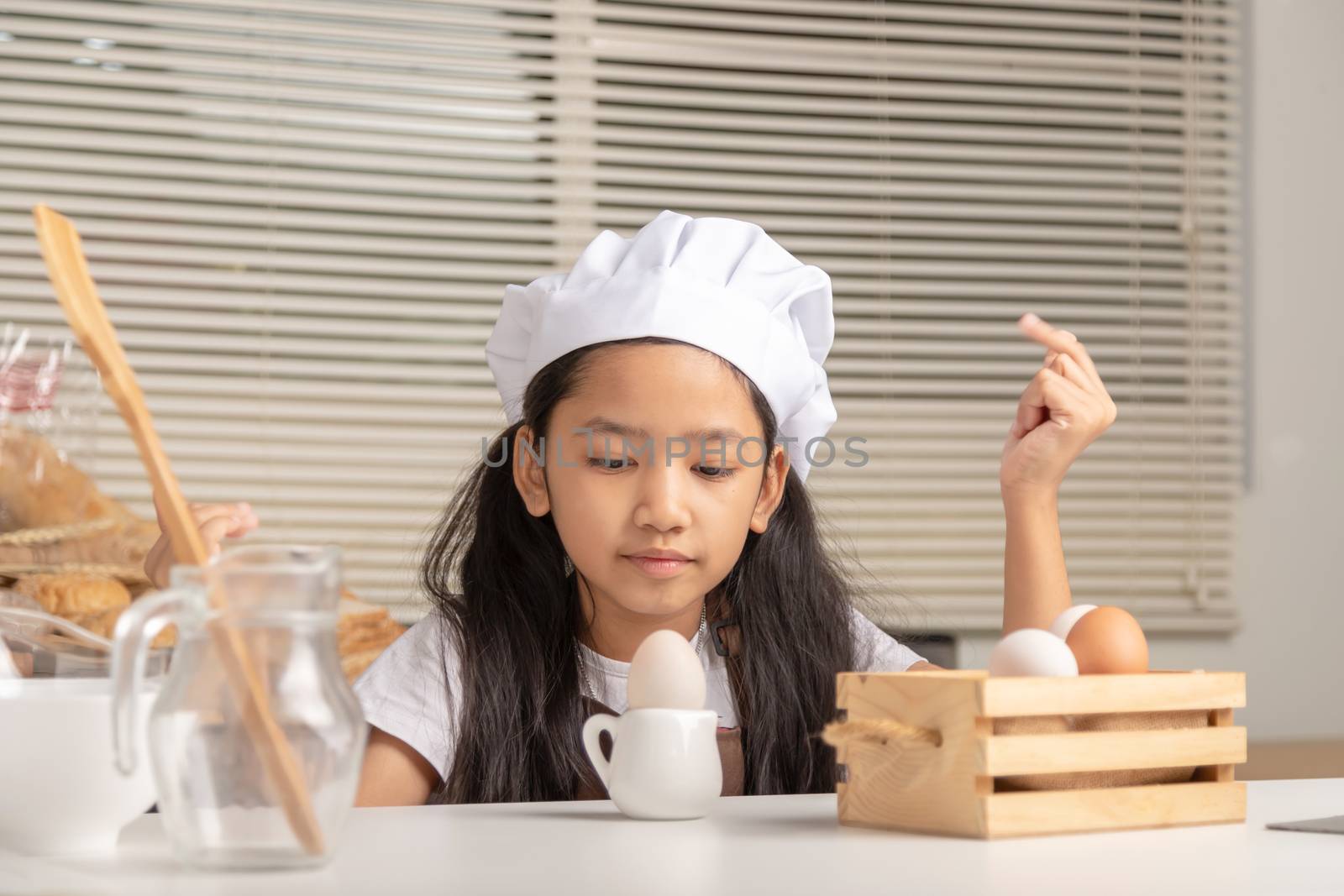 A little Asian girl wearing a white chef hat is staring at a duck egg on a small jug and puts on a white cooking table in the kitchen.