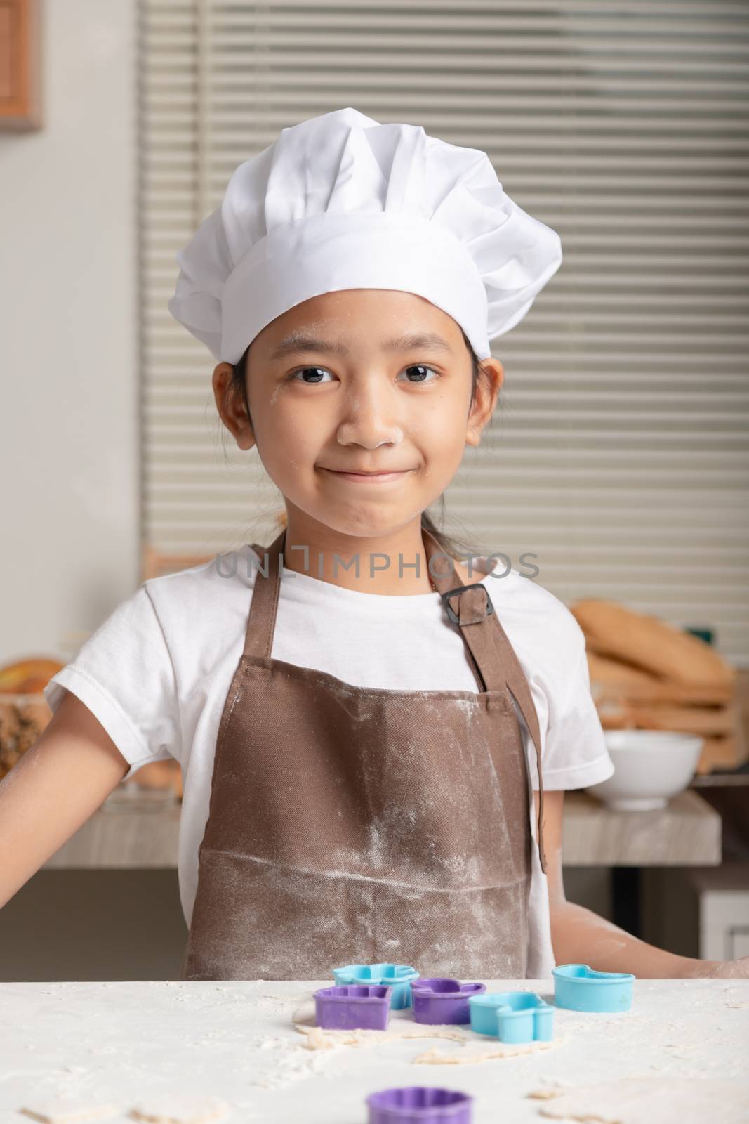 The little girl wore a white chef hat and a brown apron. The kid making homemade bakery in the kitchen. Children smiling with happiness.