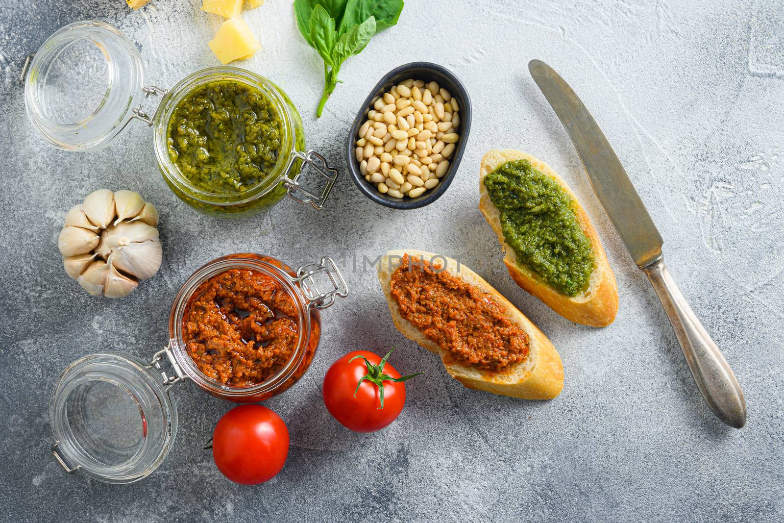 Glass jars with red and green pesto and cooking italian recipe ingredients Parmesan cheese, basil leaves, pine nuts, olive oil, garlic, salt, tomatoes breakfast panini with sauce top view on grey concrete surface space for text by Ilianesolenyi
