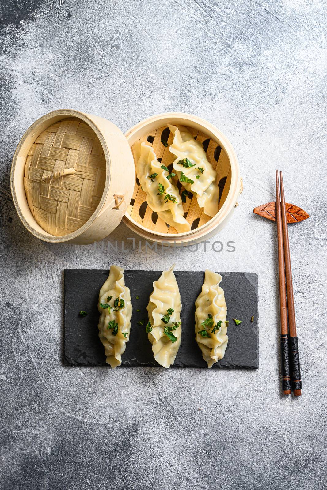 Dim sum dumplings in steamer and on a stone plate with chopsticks and fresh herbs top view on grey background by Ilianesolenyi