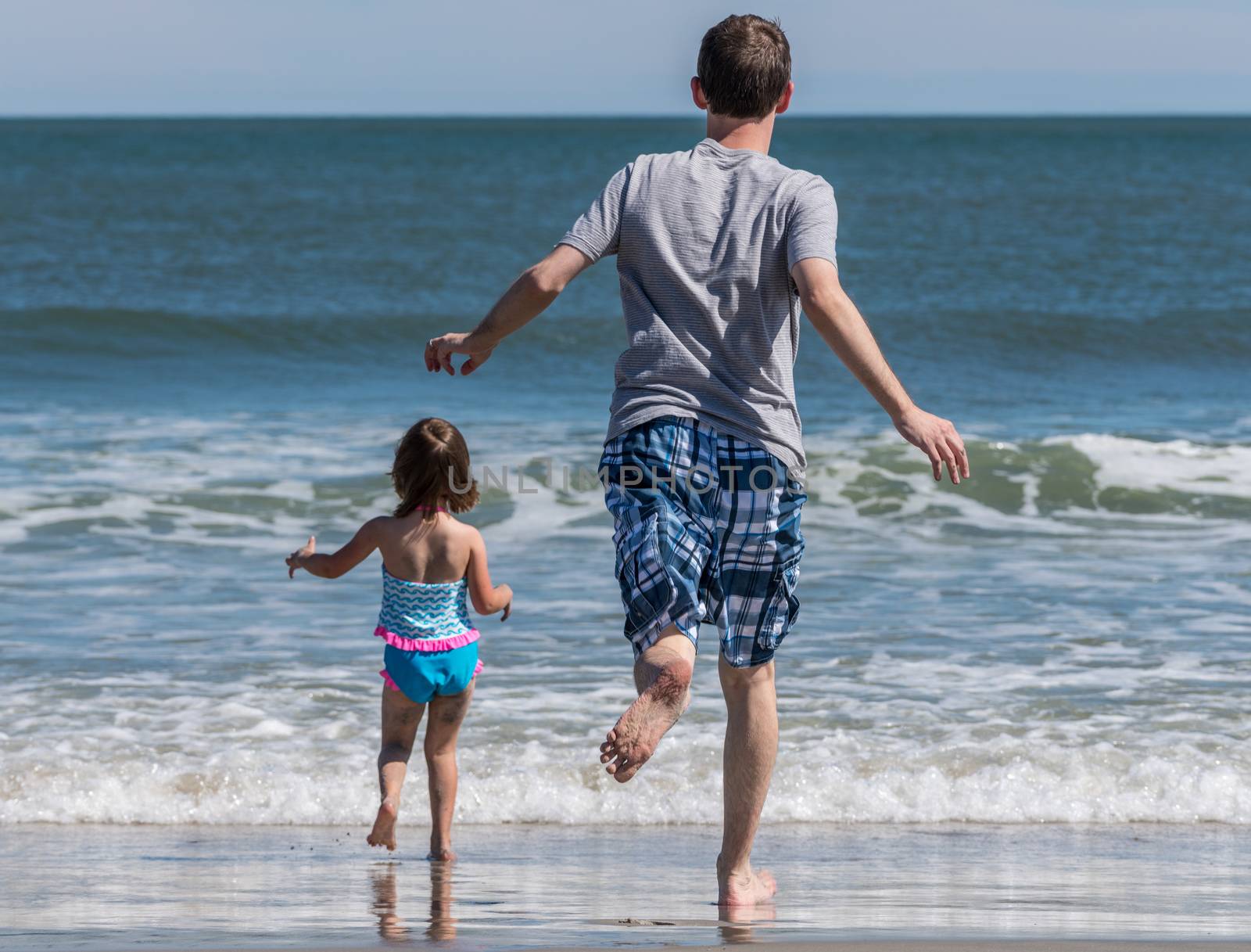 Rear view of father and young daughter running into the ocean waves by steheap