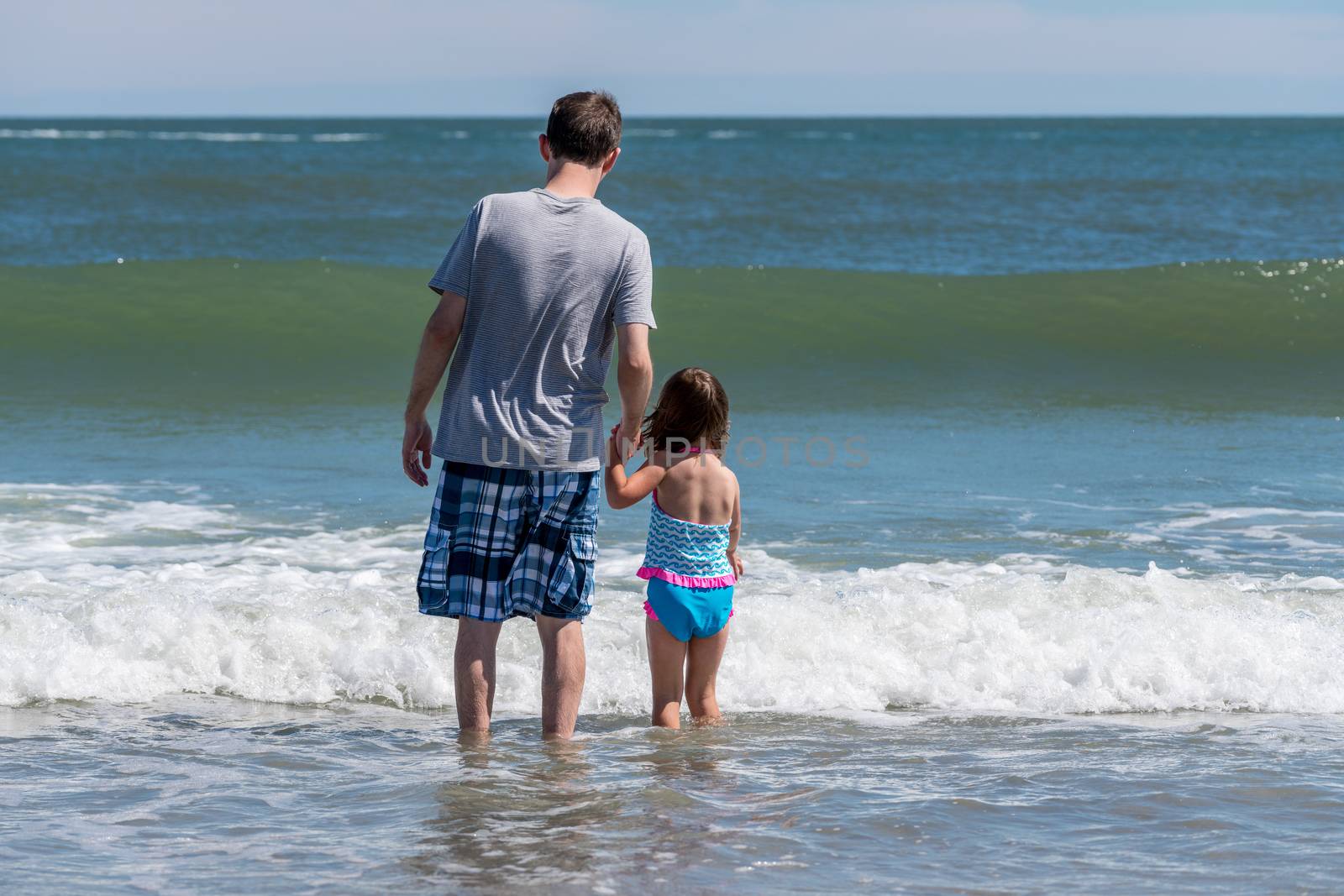 Rear view of father and young daughter facing the ocean waves by steheap