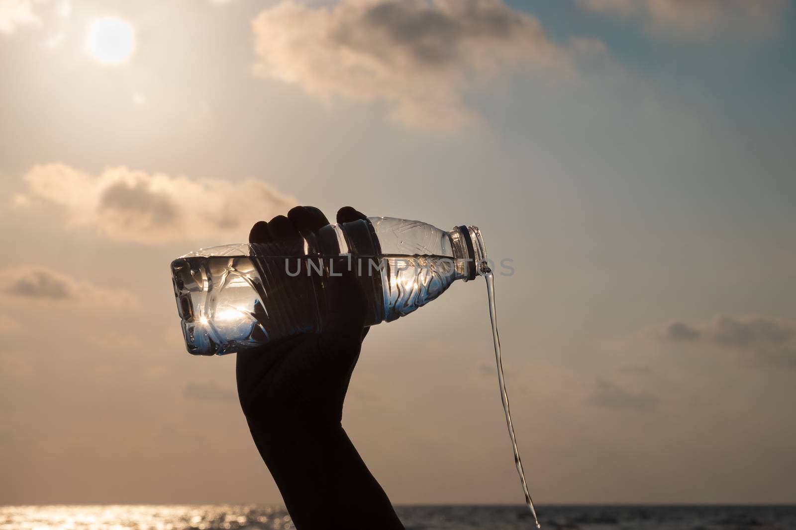 The hand is pouring water from the bottle on the beach at evenin by feelartfeelant