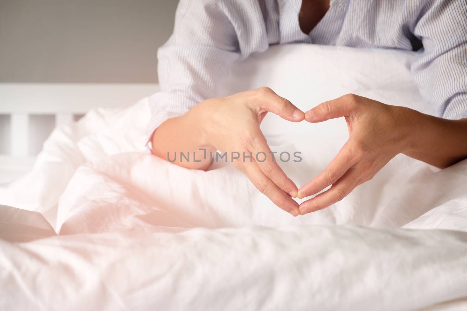 Woman is showing a gesture by hand in heart shape on the white mattress in the morning, with the orange light from the sun shining.