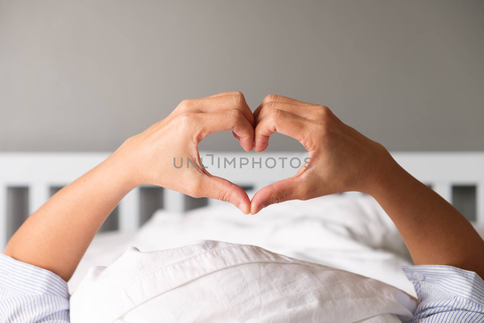 Woman is showing a gesture by hand in heart shape on the white mattress in the morning.