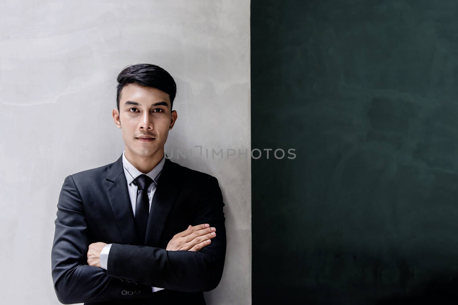 Portrait of Young Confident Businessman in Black Formal Suit. Standing by the Wall, Crossed Arms and Looking at Camera