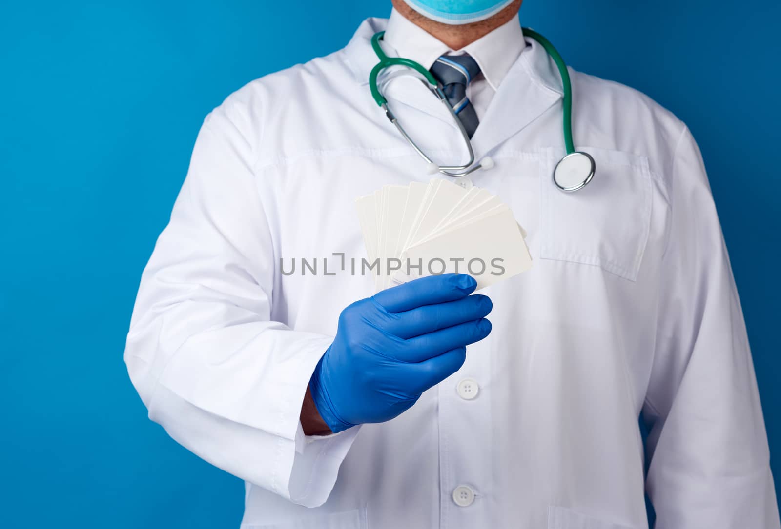doctor therapist is dressed in a white robe uniform and blue sterile gloves is standing and holding a stack of empty white paper business cards, place for inscription