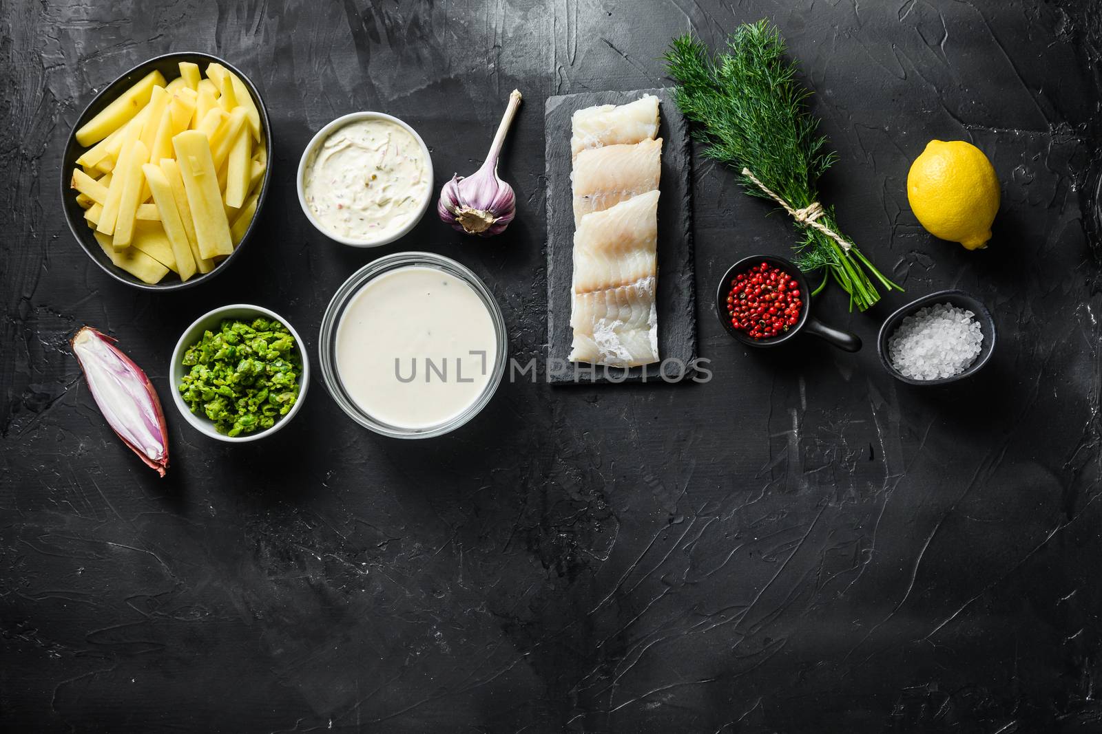 Traditional fish and chips ingredients recipe raw cod fillets on stone slate batter, potatoe, tartar sauce, minty mushy peas, lemon , shallot, mint, garlic, salt, peppercorns on black stone background concept top view space for text.