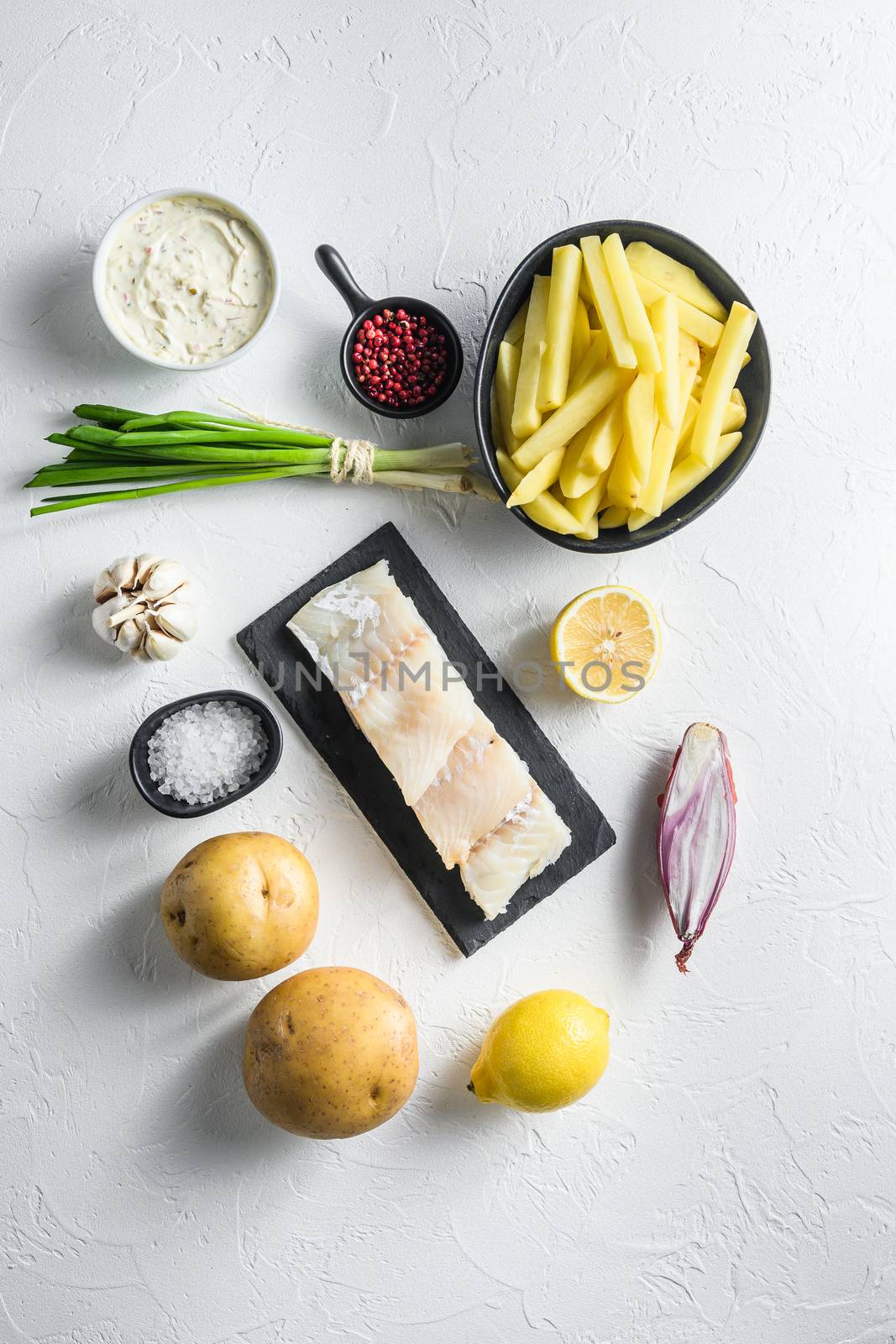 Raw cod fillet and other organic ingredients recipe fish and chips batter, potatoe, tartar sauce, lemon, capers , green herbs garlic, salt, peppercorns on white concrete textured surface or table top view by Ilianesolenyi