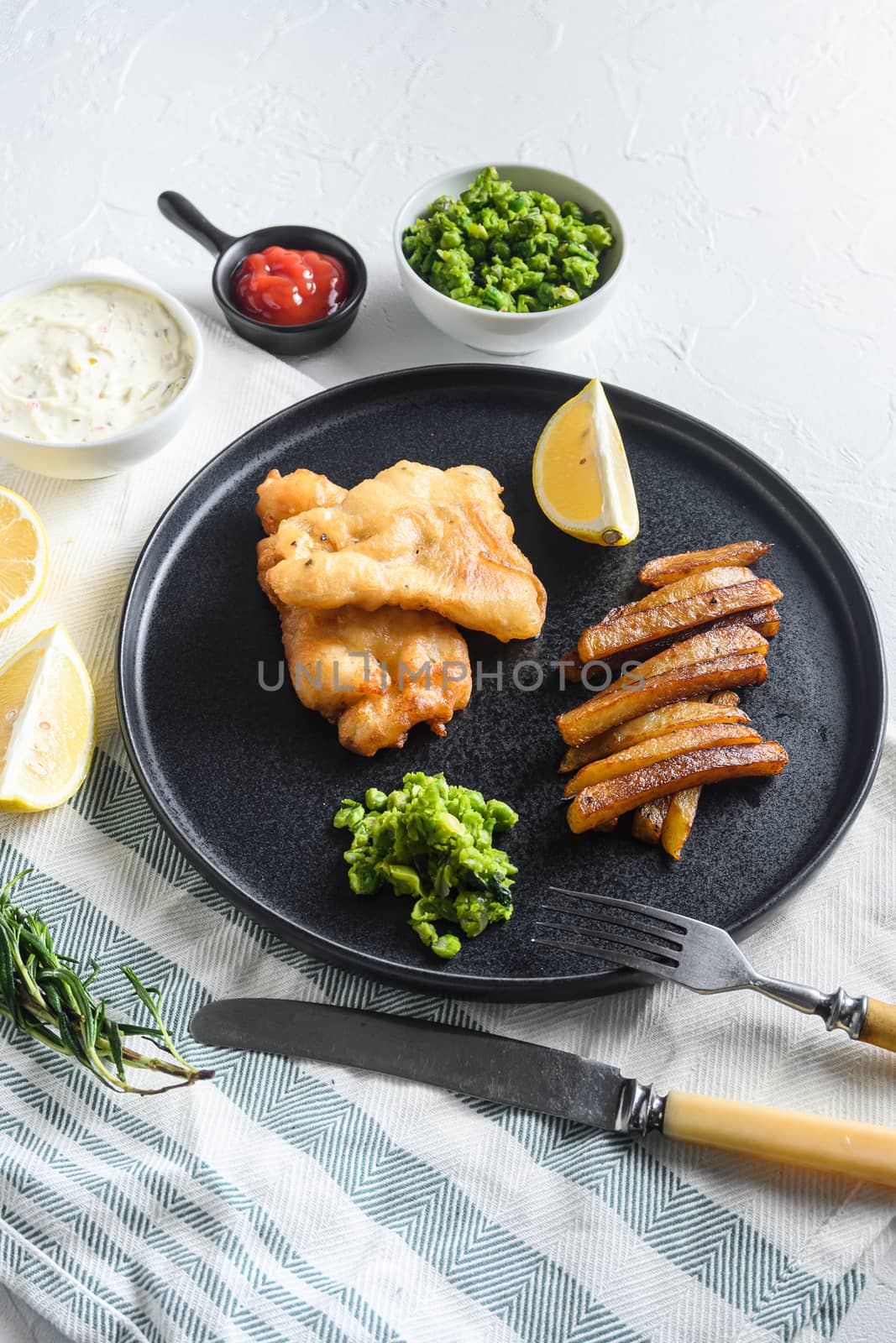 crispy fish and chips with battered cod and fries and a side of tartar sauce and mushy peas on rustic white stone table side view close-up by Ilianesolenyi