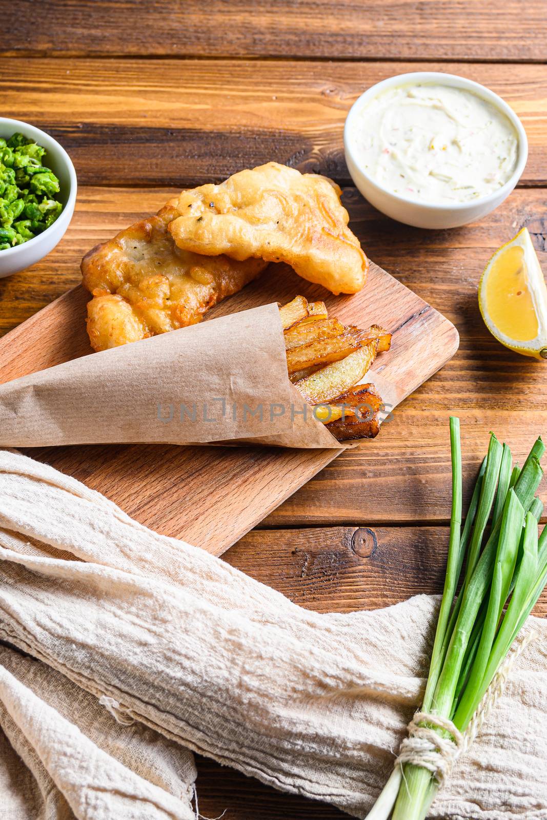 Traditional Fish and chips with mashed peas, tartar sauce inn crumpled paper cone on wood chopping board dip and lemon - fried cod, french fries, lemon slices, tartar sauce, ketchup tomatoe served in the Pub or Restaurant over light old wooden planks table vertical side view close up by Ilianesolenyi