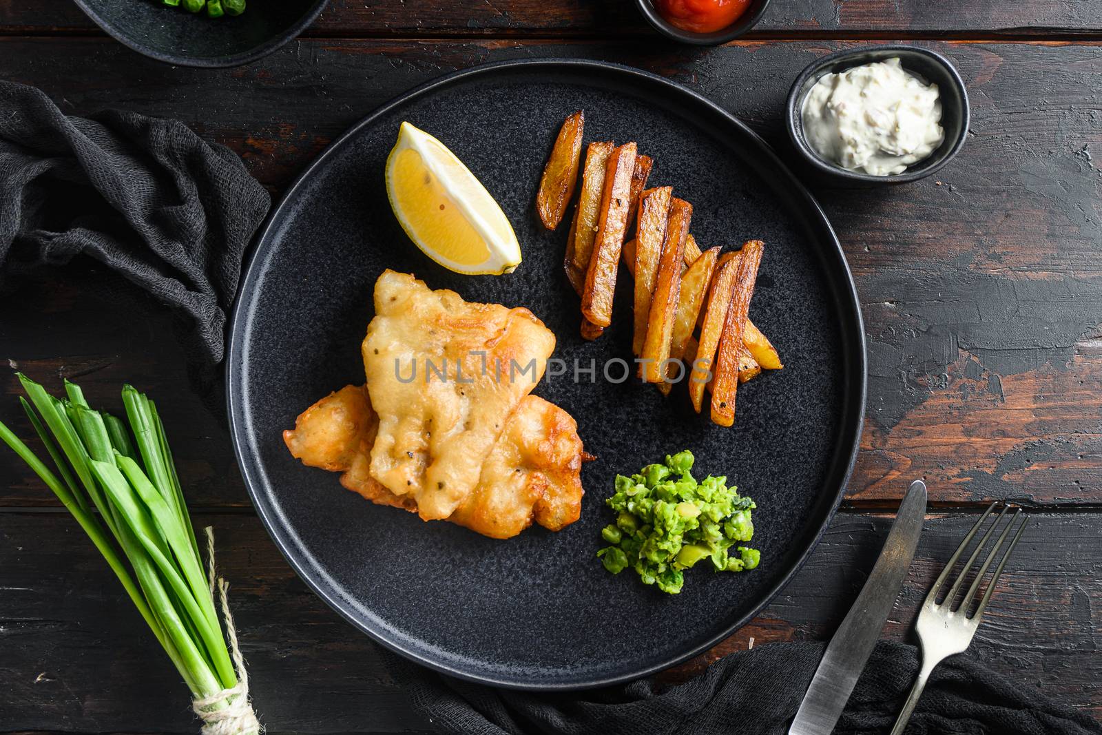 Fish and chips. Traditional british hot dish consisting of fried fish, potato chips, mushy peas and tartare sauce. served on black plate over old dark wood table in pub top view.