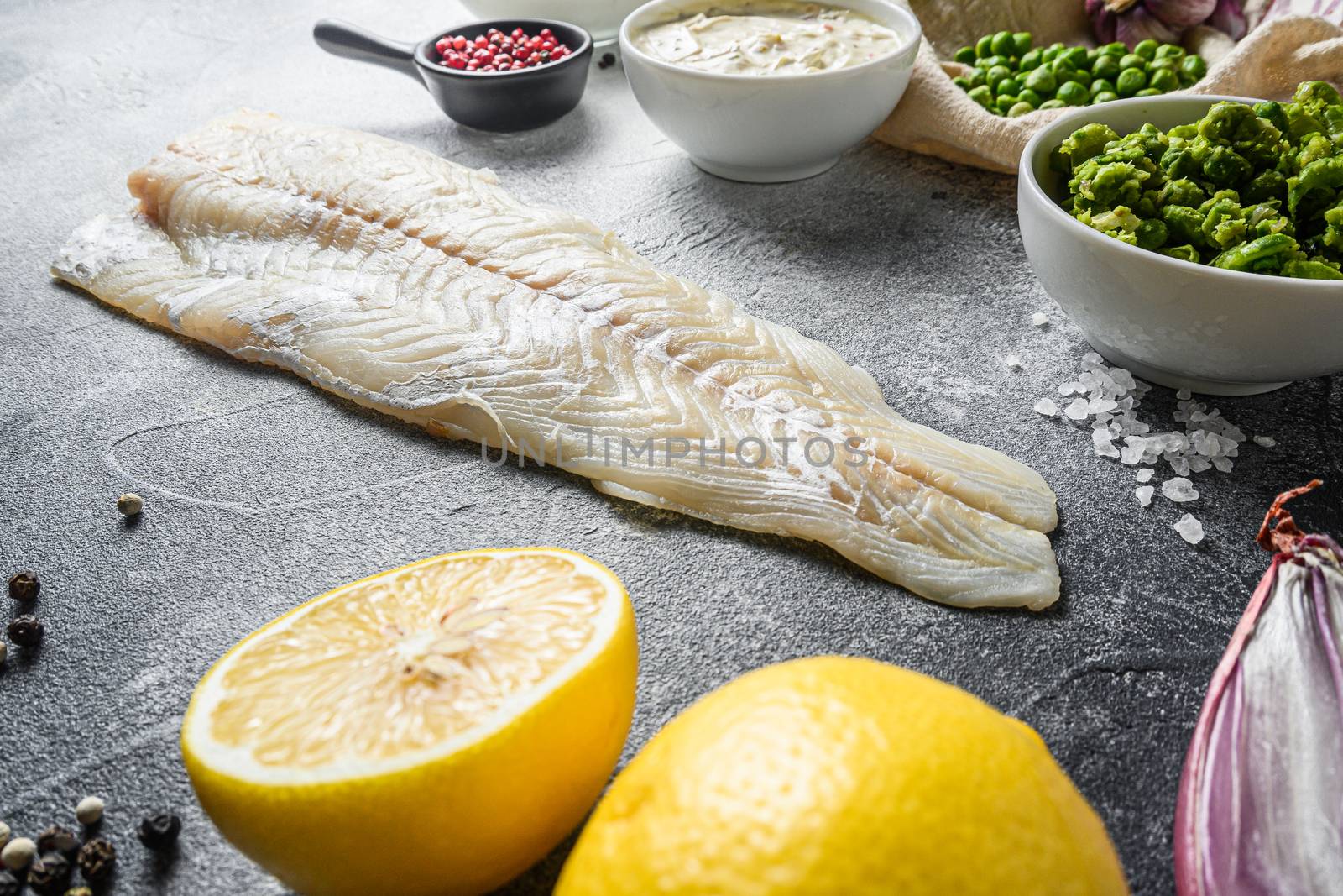 Cod fish for english traditional Fish and chips ingredients beer batter, potatoe, tartar sauce, minty mushy peas, lemon , shallot, mint, garlic, salt, peppercorns, cod fish side view grey concrete background new wide angel