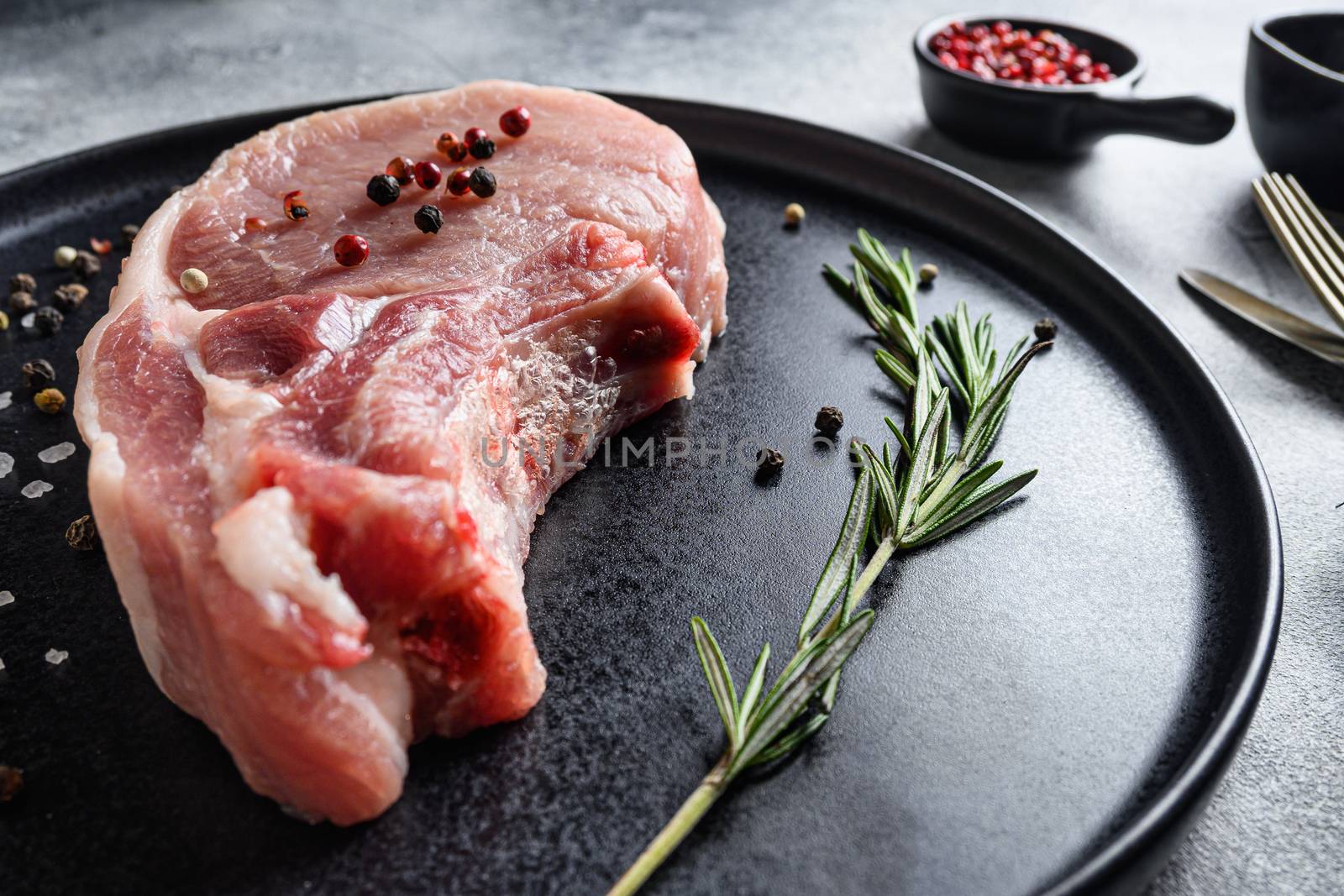 Raw Pork Loin chops in a black round plate on a grey textured stone background with rosemary garlic peppercorns ingredients for grill close up selective focus new wide angel by Ilianesolenyi
