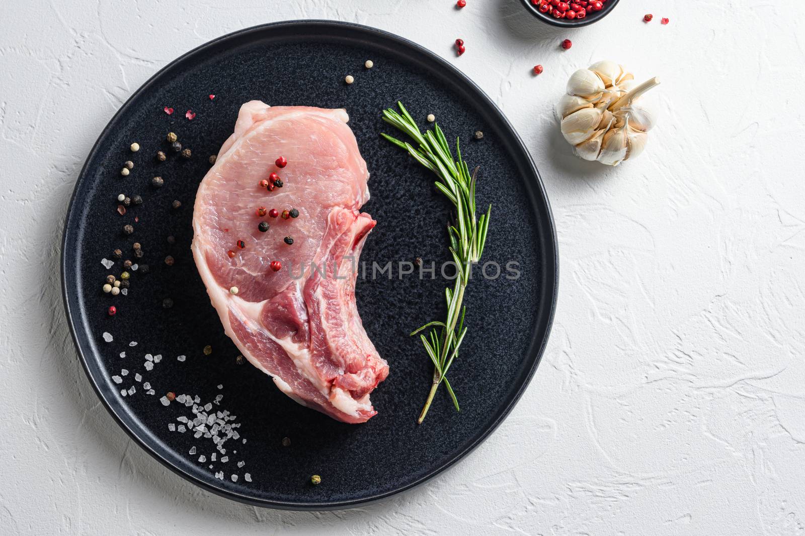 Fresh pork steak in a black round plate on a white stone background with rosemary garlic peppercorns ingredients for grill top view space for text by Ilianesolenyi