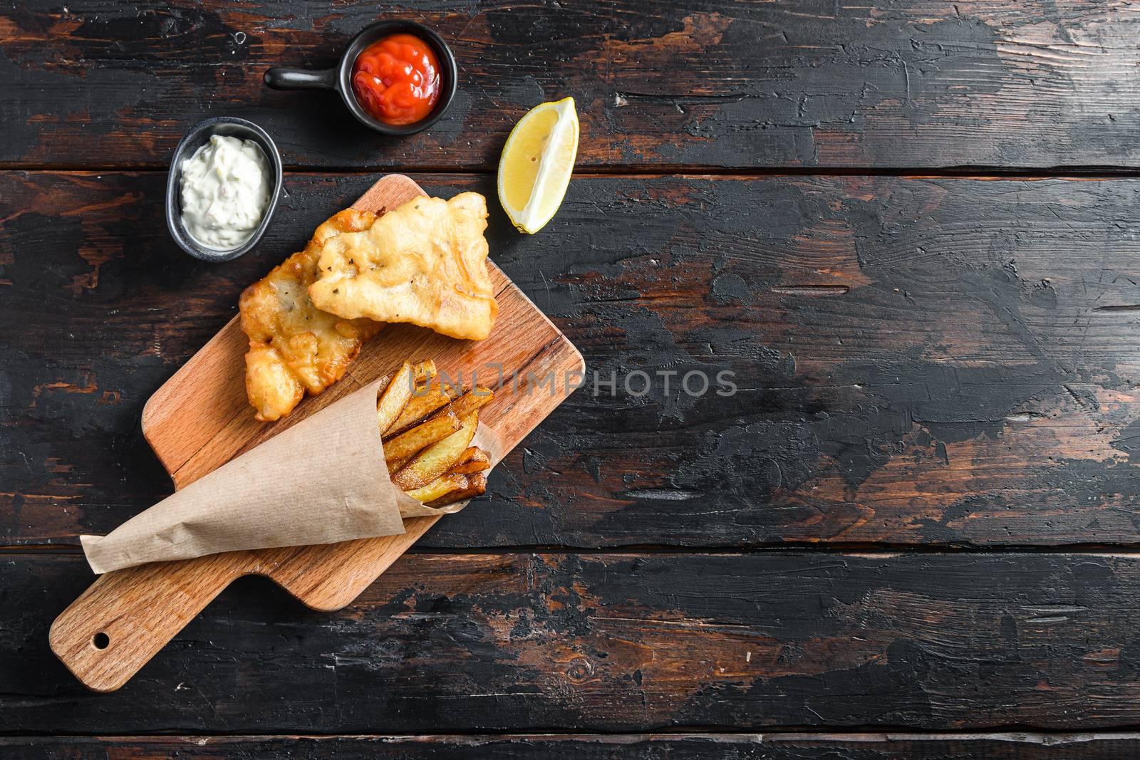 Fish and chips in a paper cone on wood chopping board dip and lemon - fried cod, french fries, lemon slices, tartar sauce, ketchup tomatoe and with mushy peas served in the Pub or Restaurant over old wooden planks dark table top view space for text or recipe