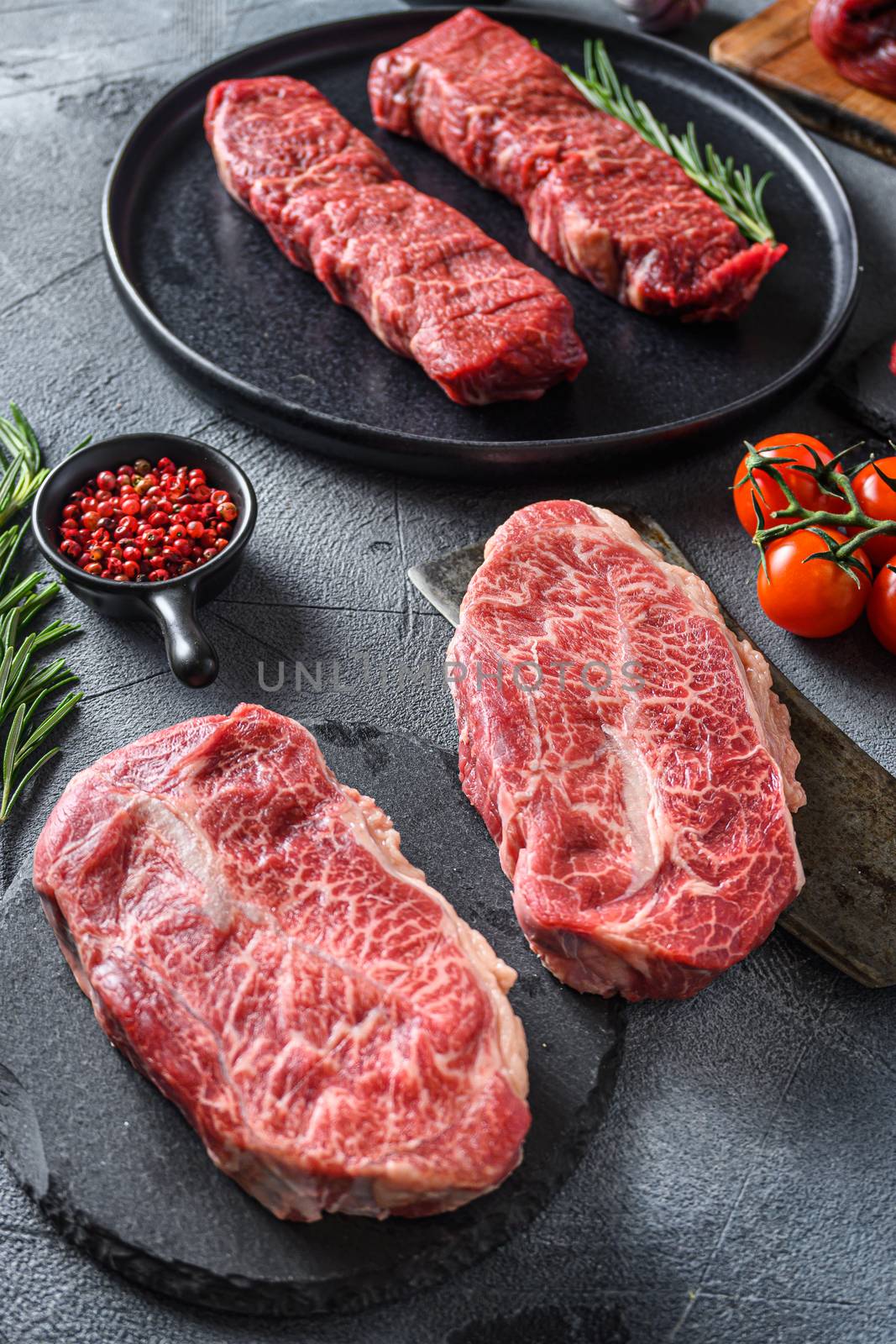 Organic feather steak steak on chopping board, marbled beef over other alternative cuts with herbs tomatoes peppercorns over grey stone surface background side view close up selective focus new wide angle.